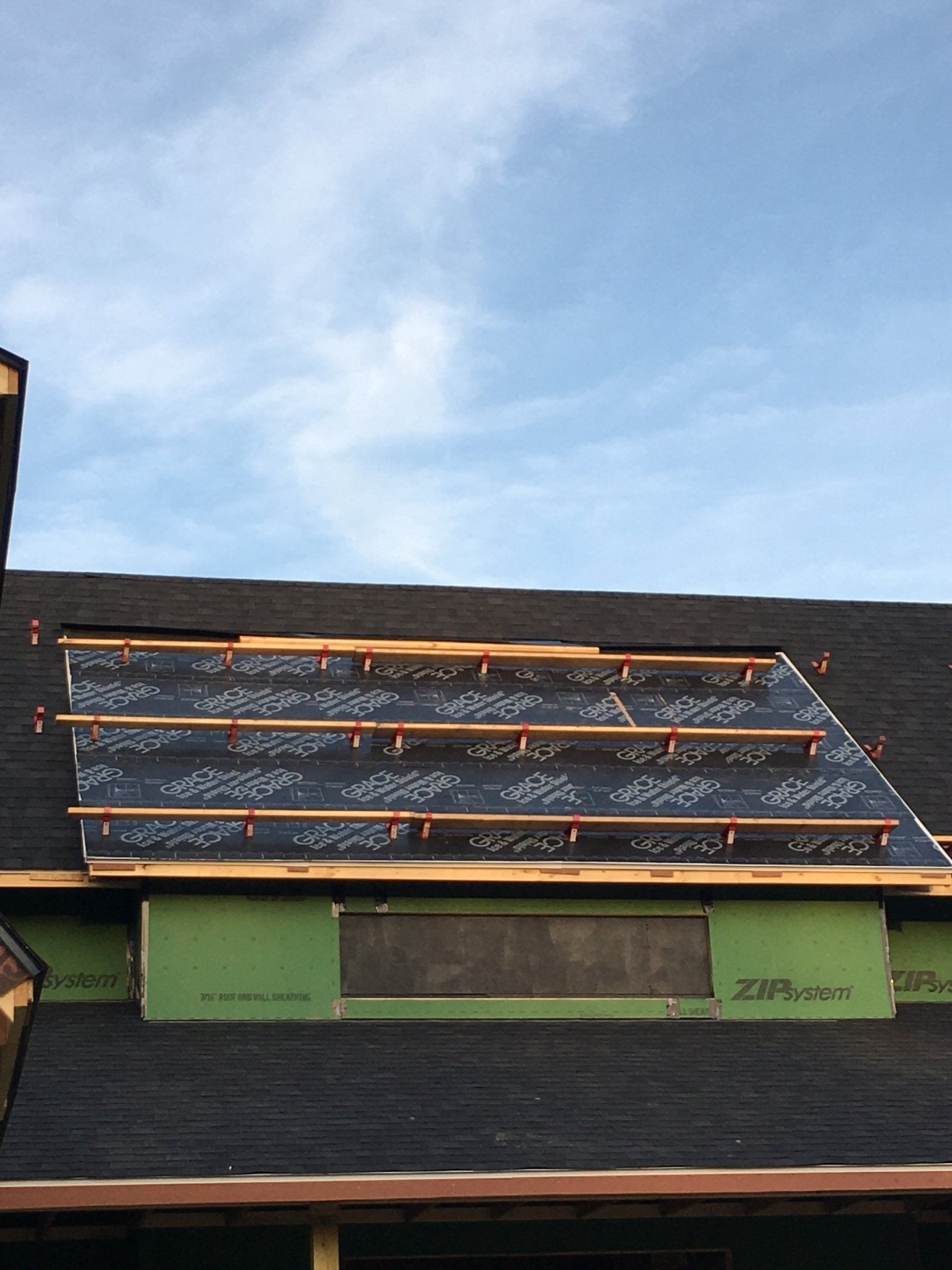 Roof under construction with black shingles, orange battens, and green sheathing against a blue sky