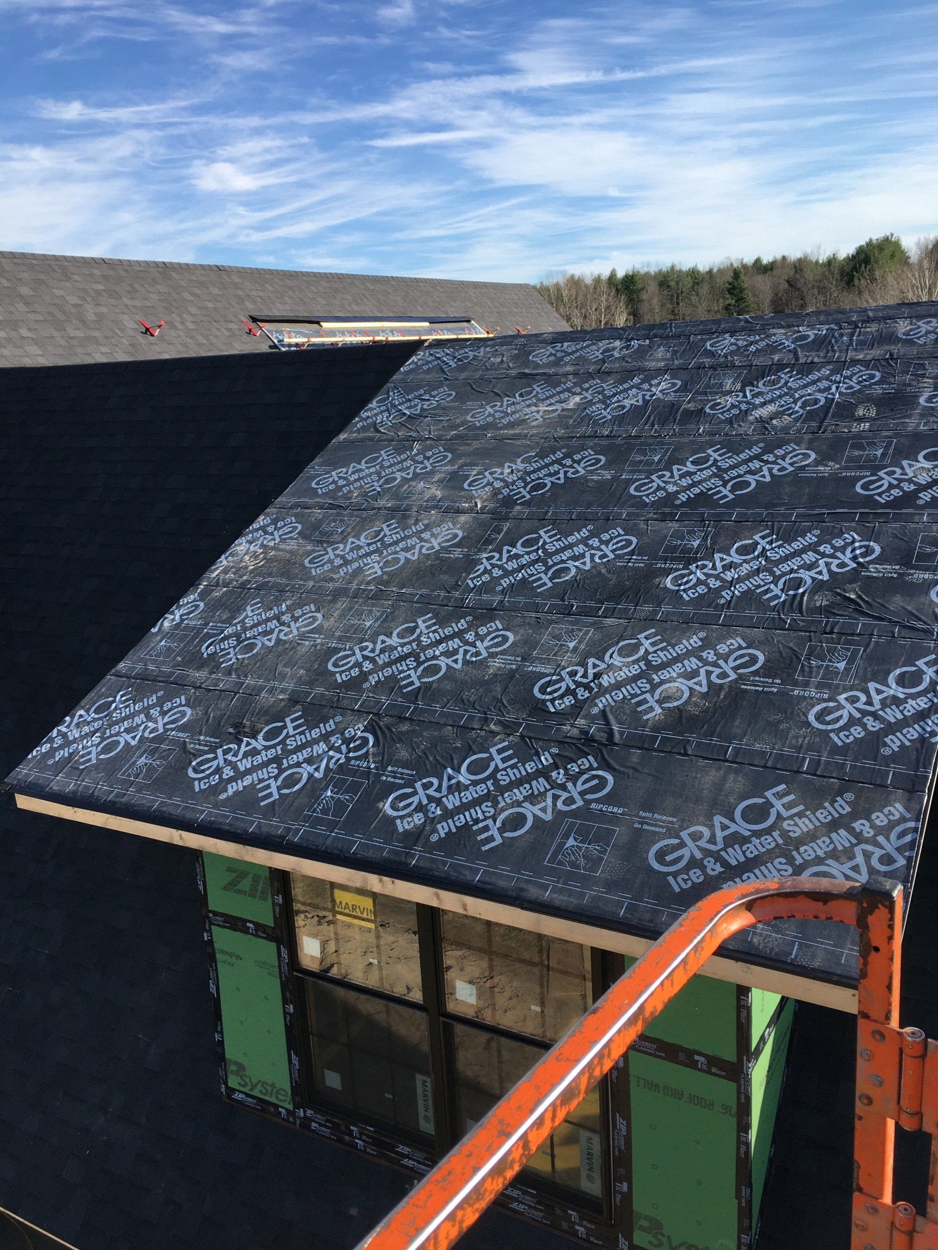 Roof under construction with black underlayment, green scaffolding, and an orange ladder against a blue sky