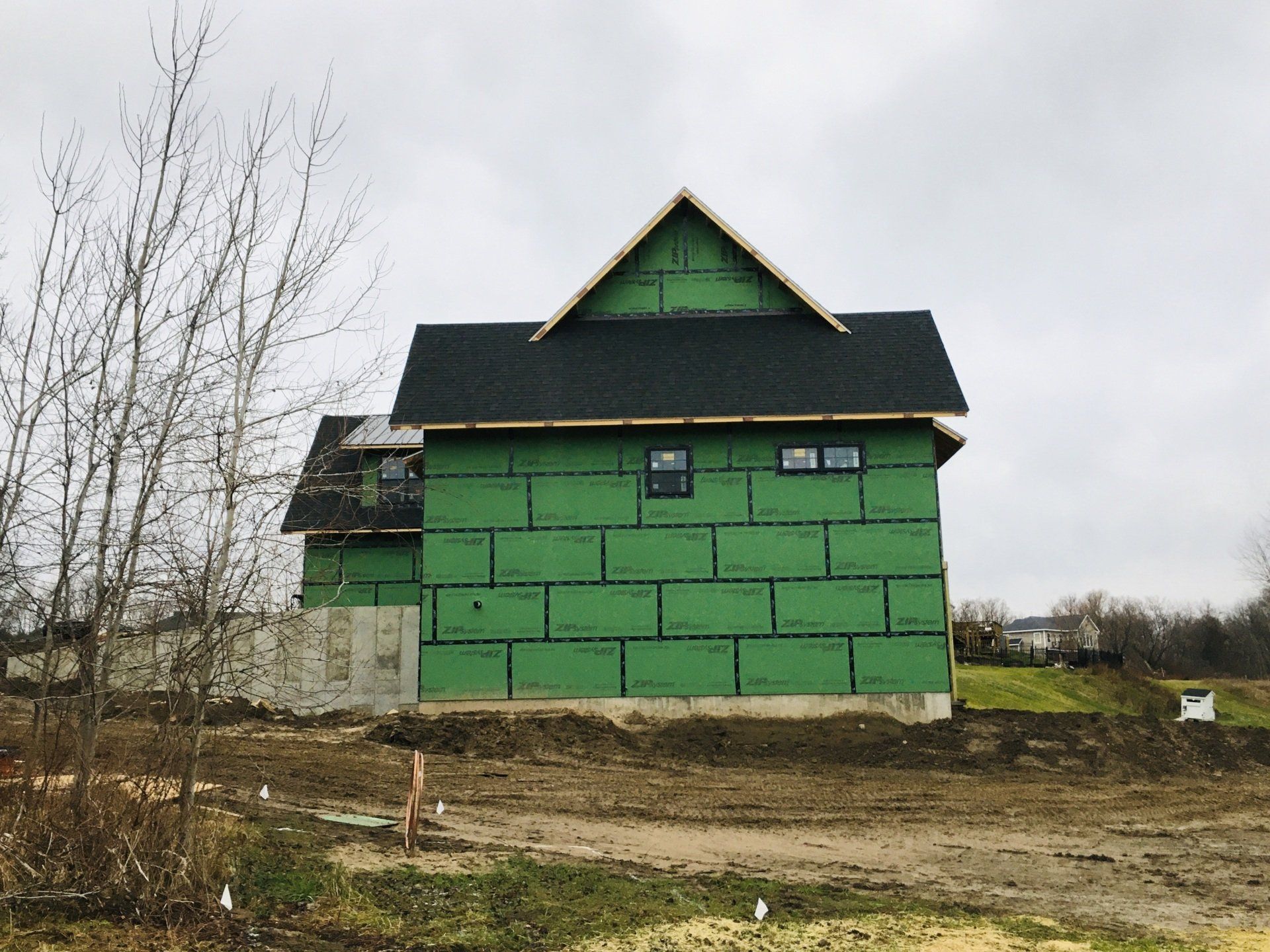 Green house under construction with a dark roof beside a muddy lot and bare trees
