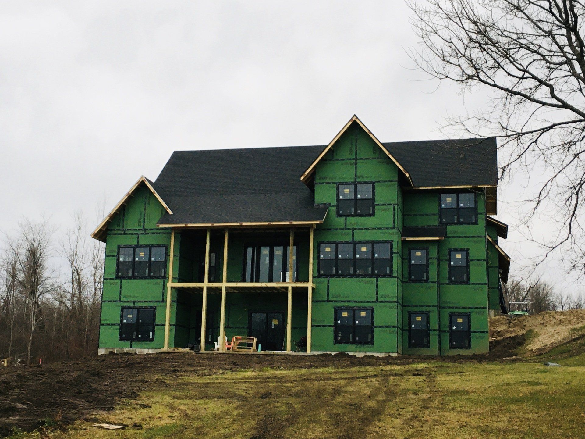 Green two-story house under construction with black roof on a grassy hillside, bare trees in the background