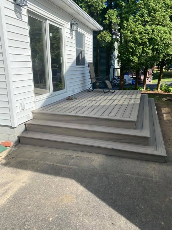 White house side deck with steps, railing, and chairs beside a driveway and trees