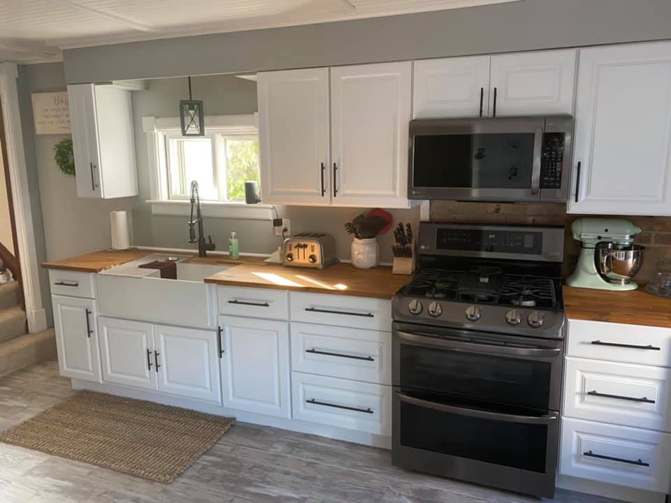 Bright white kitchen with wood countertops, black stove and microwave, sink, cabinets, and woven rug.