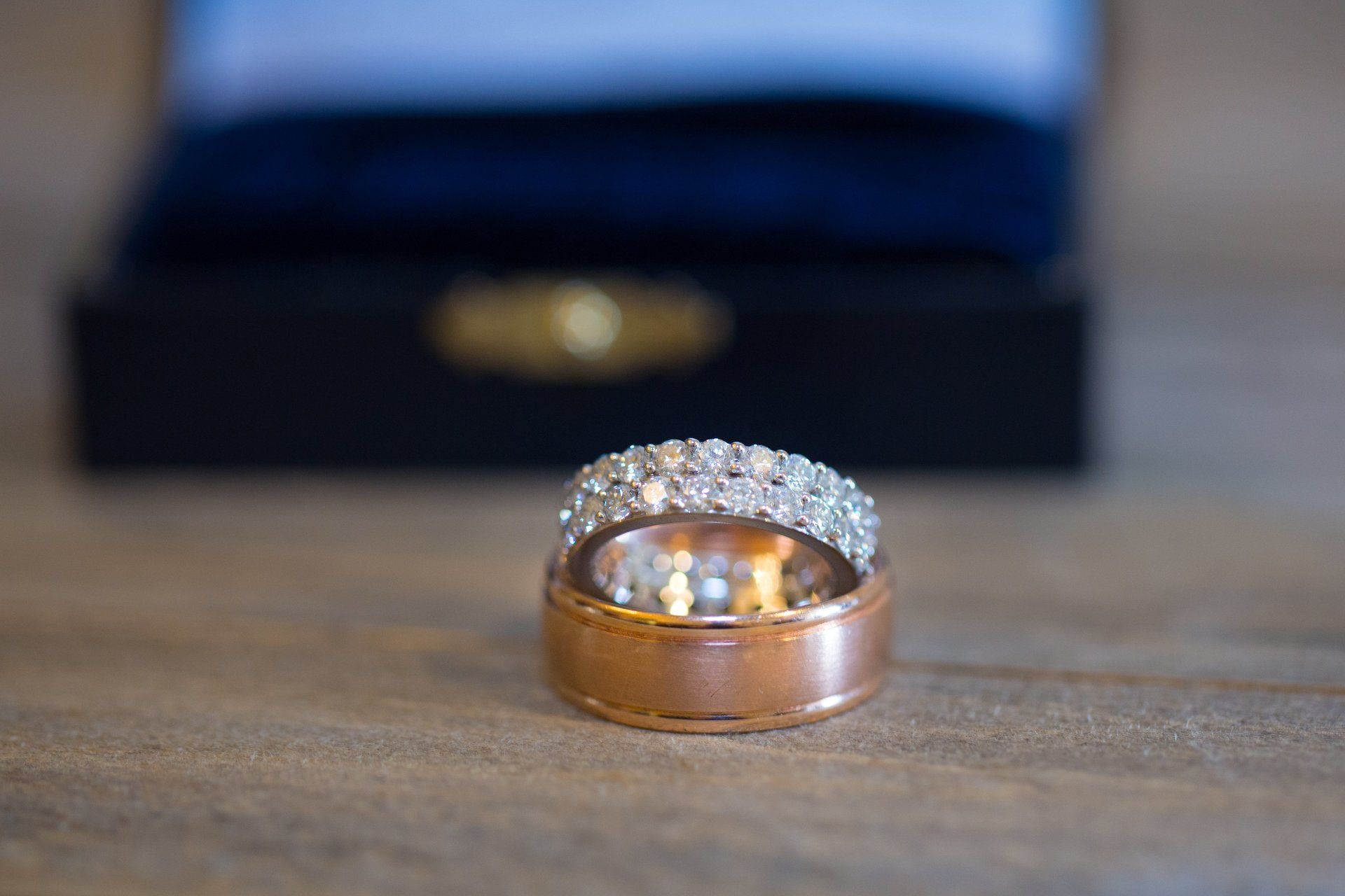Two wedding rings stacked, one with diamonds, in front of a blue velvet ring box on wood.