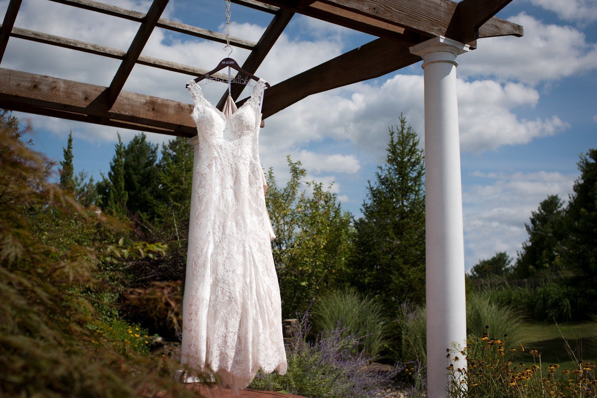 Wedding dress hanging on a wooden arbor with a white column, trees, and sky.