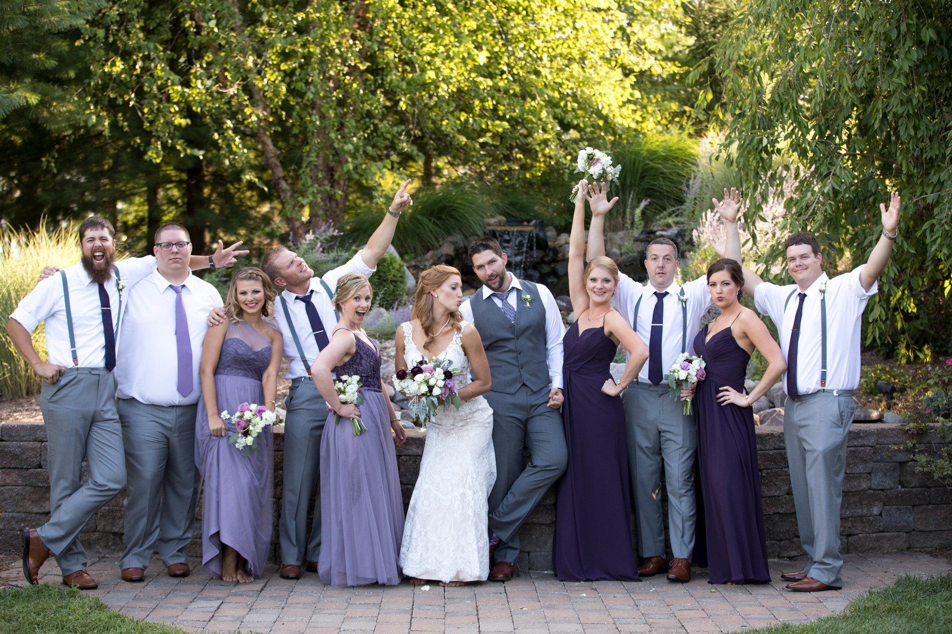 Wedding party poses outdoors. Bride and groom stand with bridesmaids and groomsmen in a garden; some raise arms.