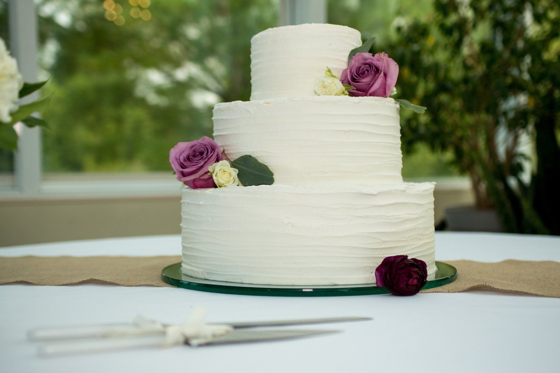Three-tiered white wedding cake decorated with purple roses on a table, knife set in foreground.