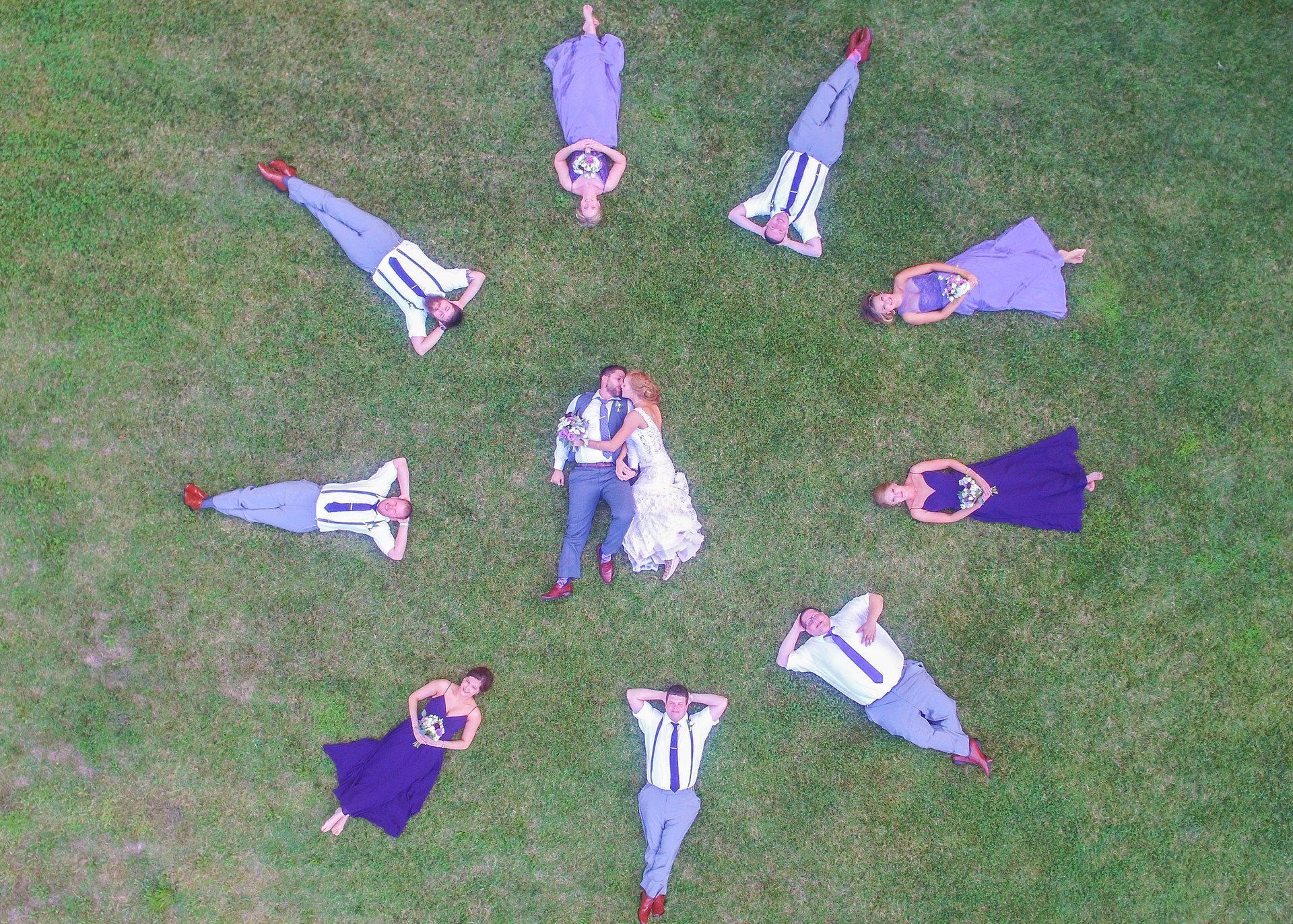 Wedding party lying in a circle around a kissing bride and groom on a grassy field.
