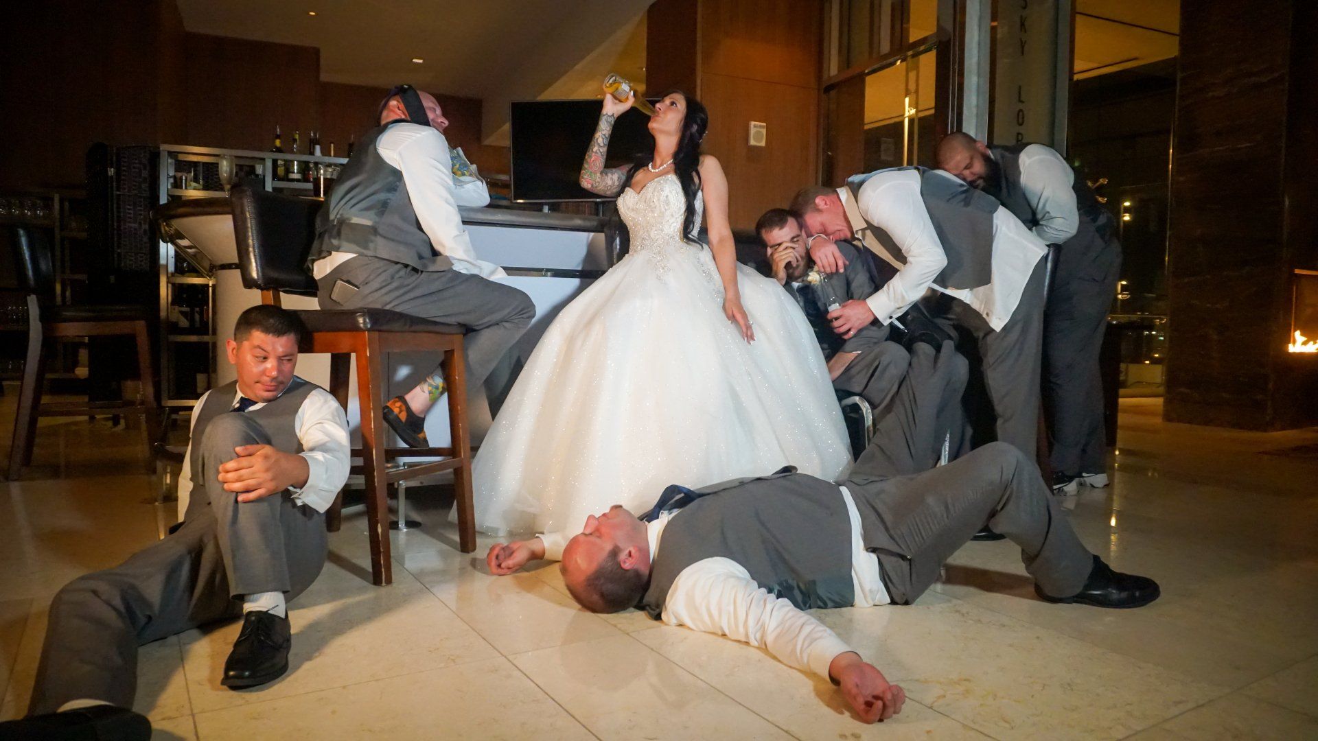 Bride and groomsmen pose comically in formal wear. Some are on the floor, seemingly drunk, while the bride drinks from a glass.