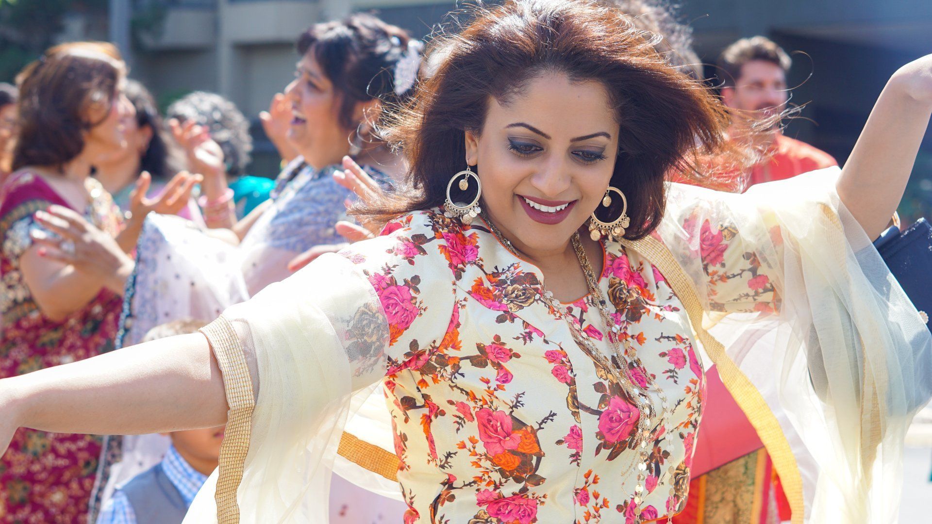 Woman dancing joyfully outdoors, wearing floral top, gold earrings, smiling. Others in background.
