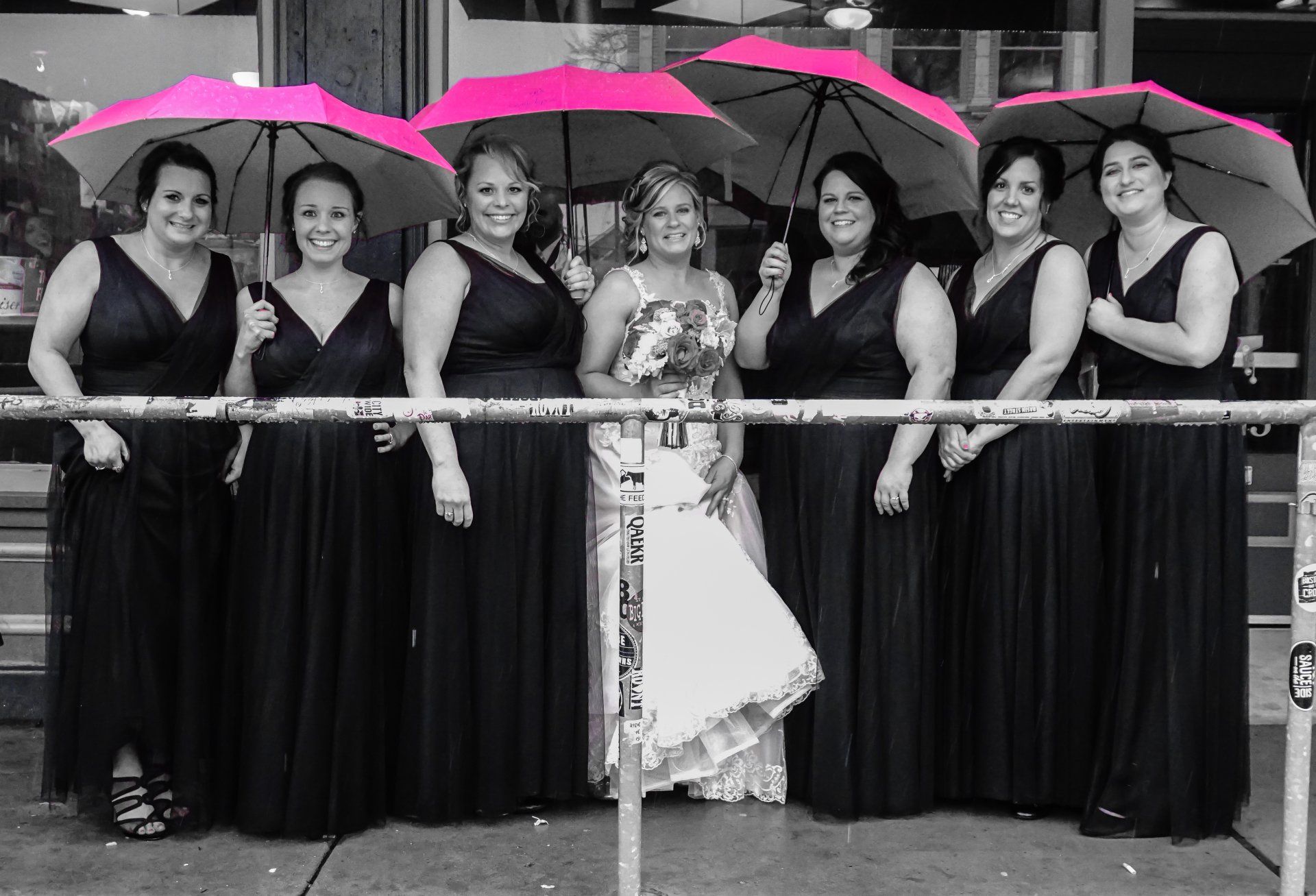Bridesmaids and bride pose with pink umbrellas; black dresses, outdoor setting, black and white except umbrellas.