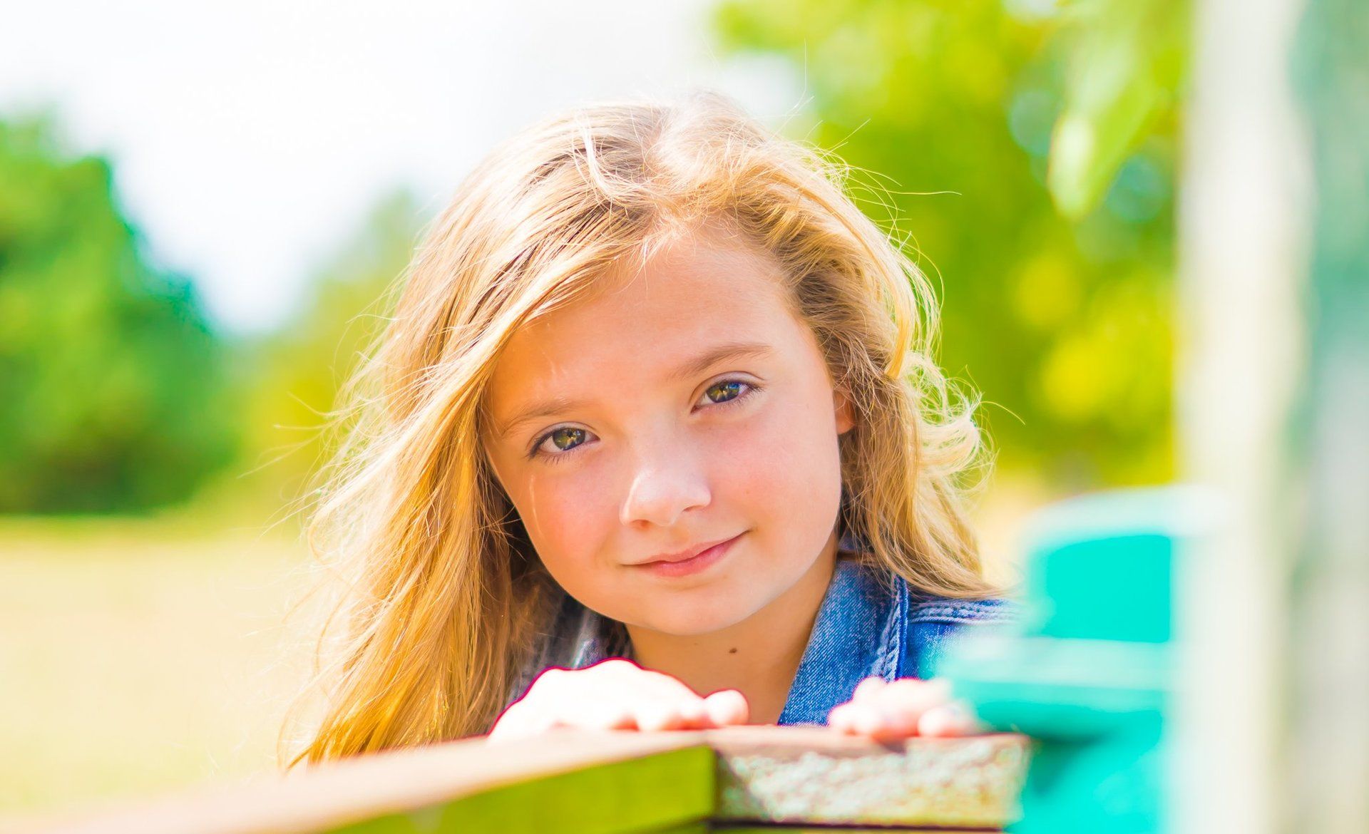 Blond-haired child peeks over a wooden fence, smiling, against a blurred green and blue background.