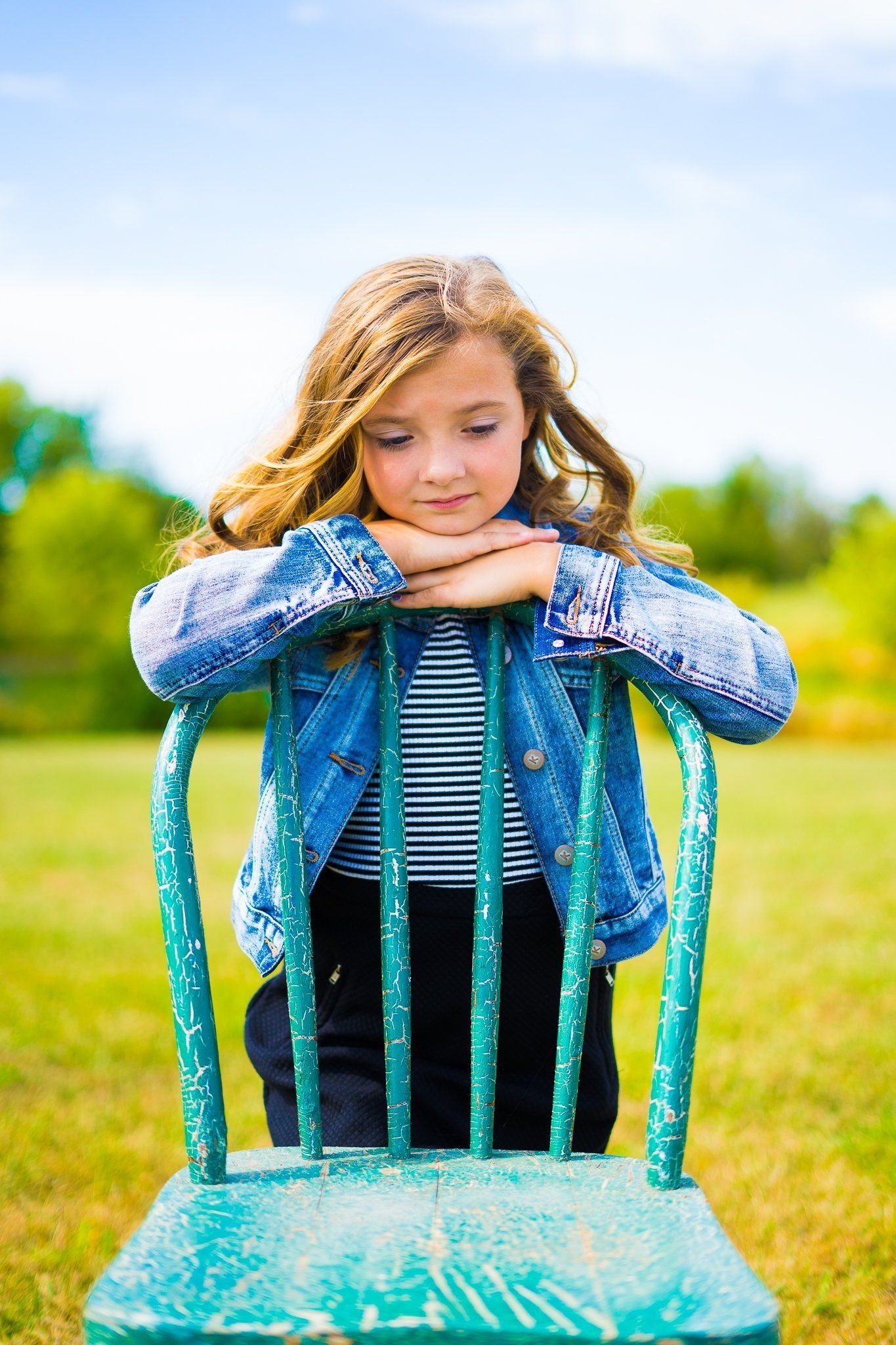 Girl in denim jacket rests chin on back of teal chair in grassy field.