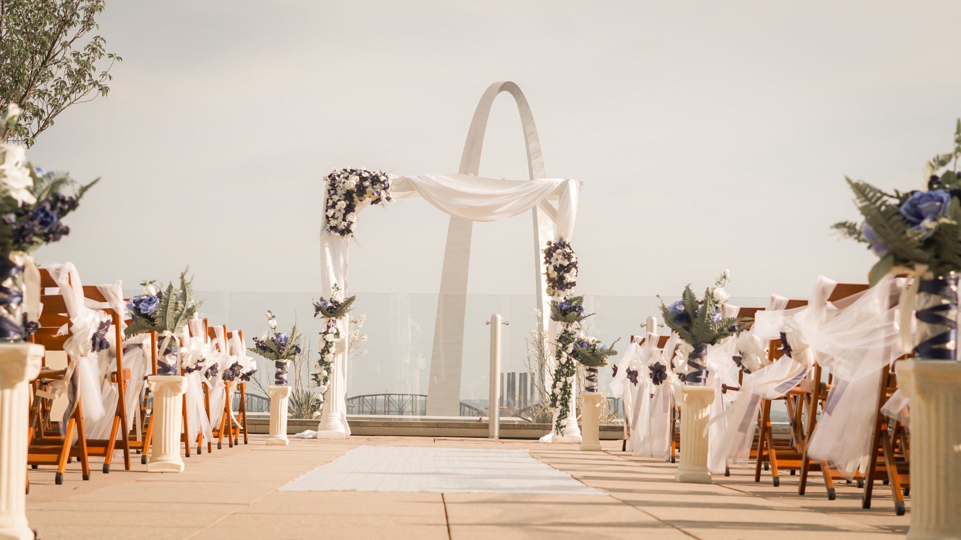 Wedding ceremony setup, arch with draping and flowers, chairs line a walkway, city skyline backdrop.