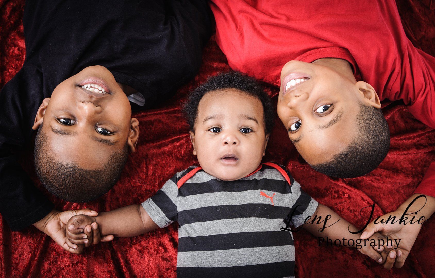 Three children lying on red blanket, smiling. Older boys in red and black shirts, holding baby's hands.