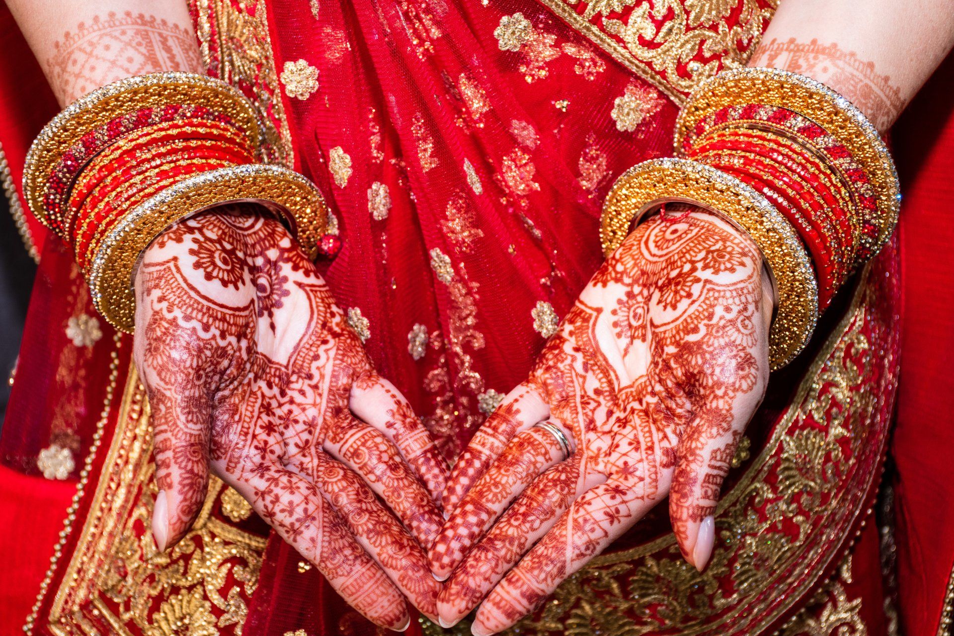 Hands with henna tattoos, wearing gold bangles, forming a heart in front of a red sari.