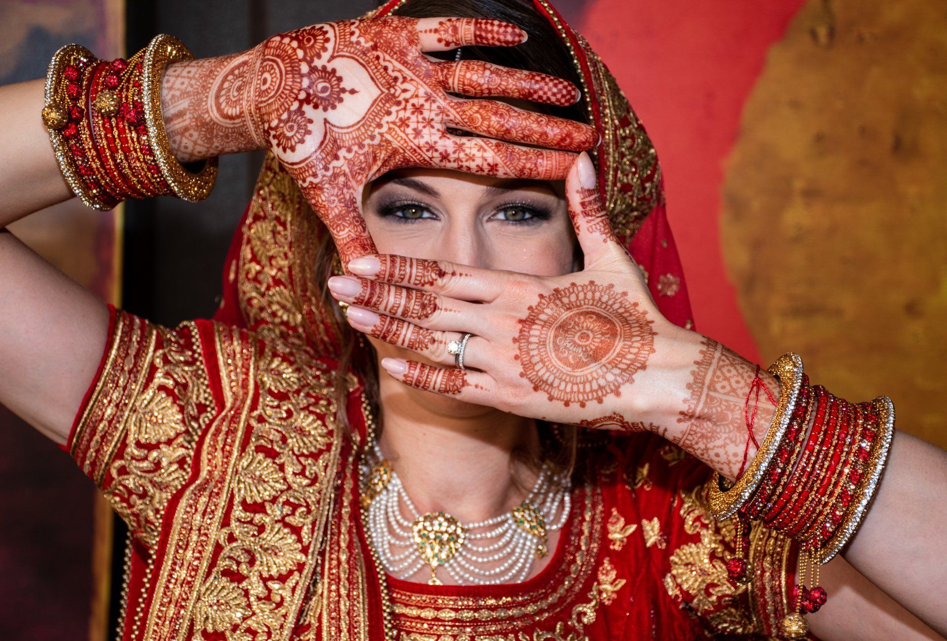 Woman wearing ornate red and gold Indian bridal attire, hands framing face, henna designs visible.