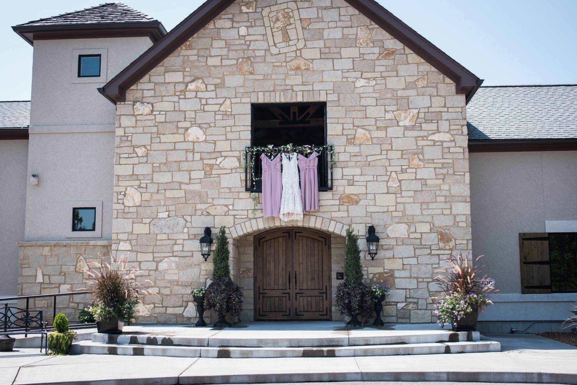 Stone building with wooden doors, a balcony with three light-colored dresses hanging, and potted plants.