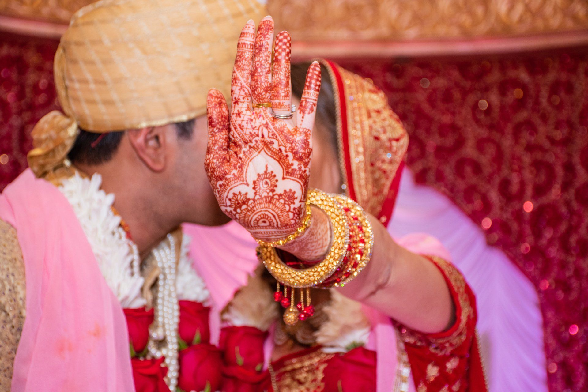 Bride in red and gold sari blocks kiss with hennaed hand, groom in turban. Wedding.