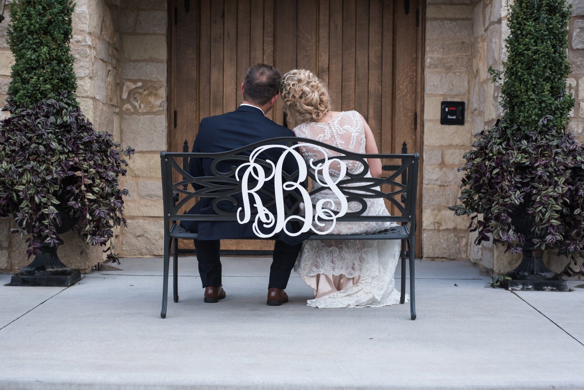 Newlyweds sit on a bench with monogram, facing away. Beige stone building with wooden door, potted plants.