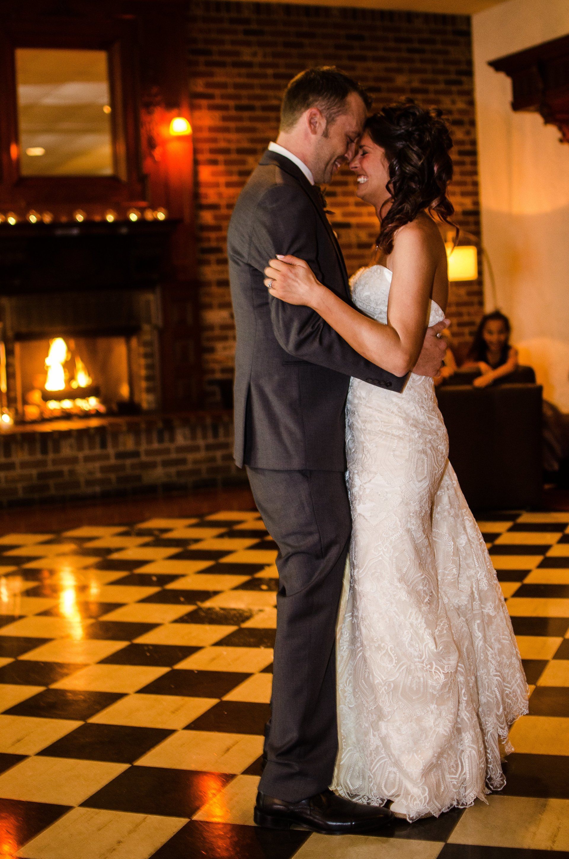 Bride and groom dance closely, forehead to forehead, near fireplace; black and white checkered floor.