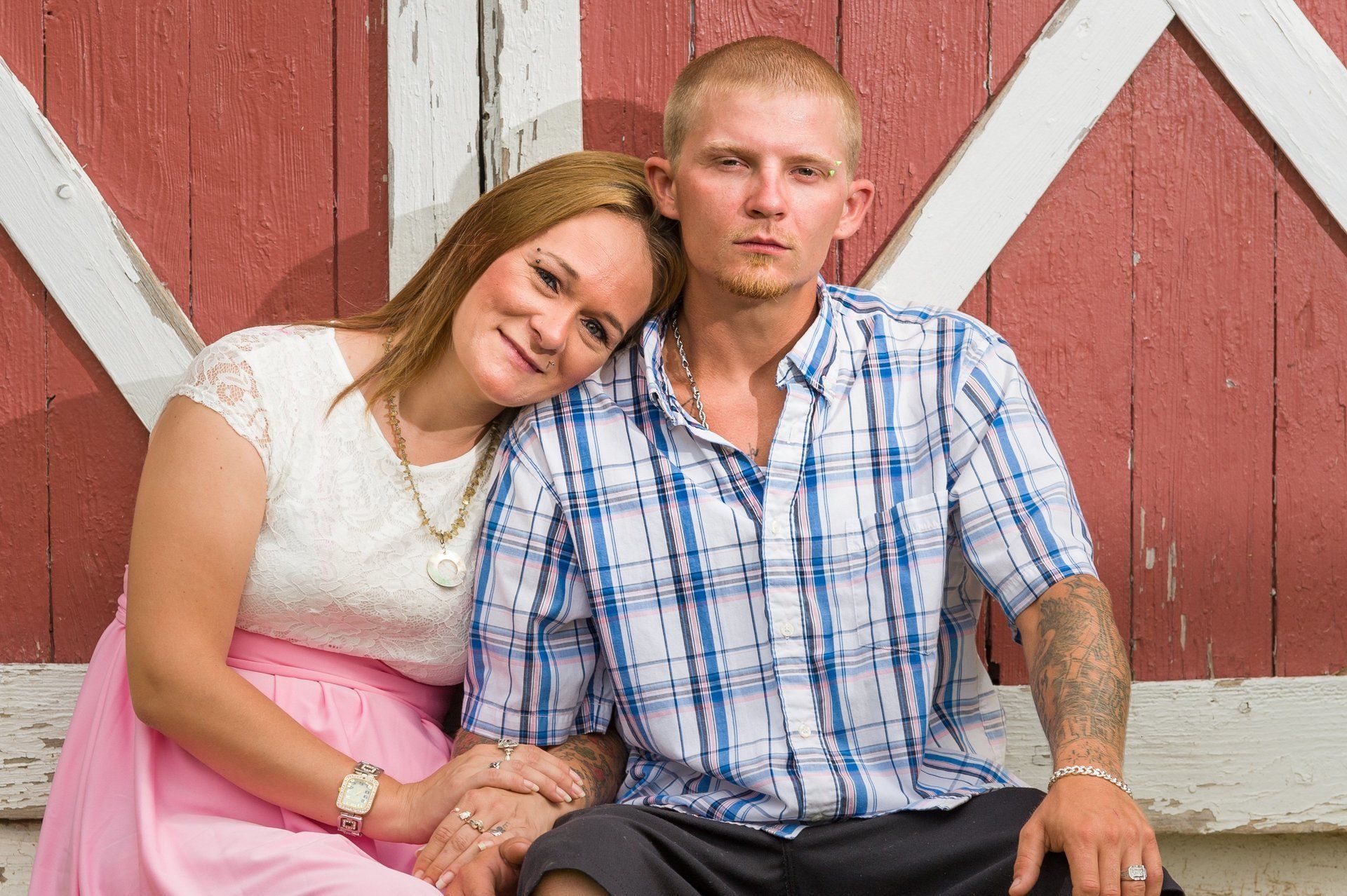 Couple sitting, leaning against a red barn. Woman in pink skirt, man in plaid shirt, both holding hands.