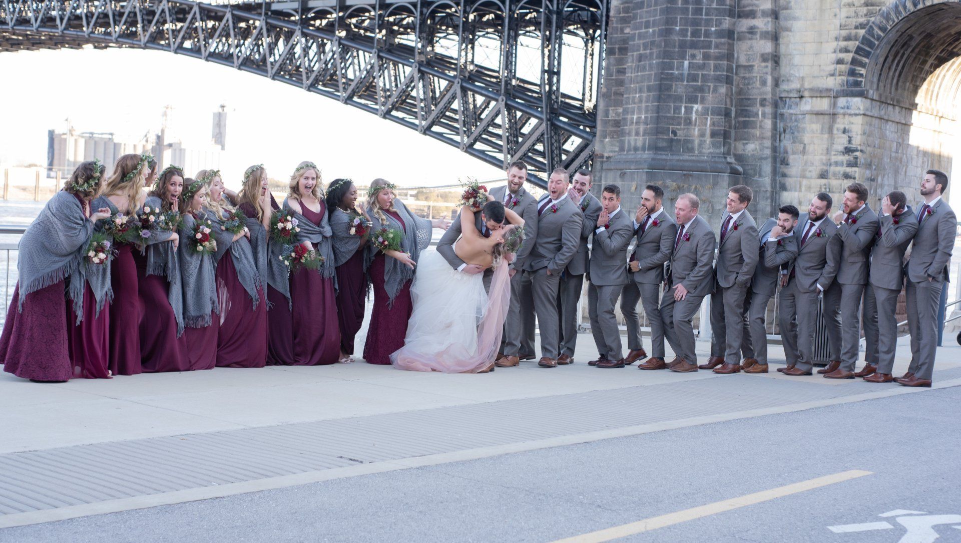 Wedding party group photo under a bridge. Bride and groom embrace, surrounded by bridesmaids in burgundy dresses and groomsmen in grey suits.