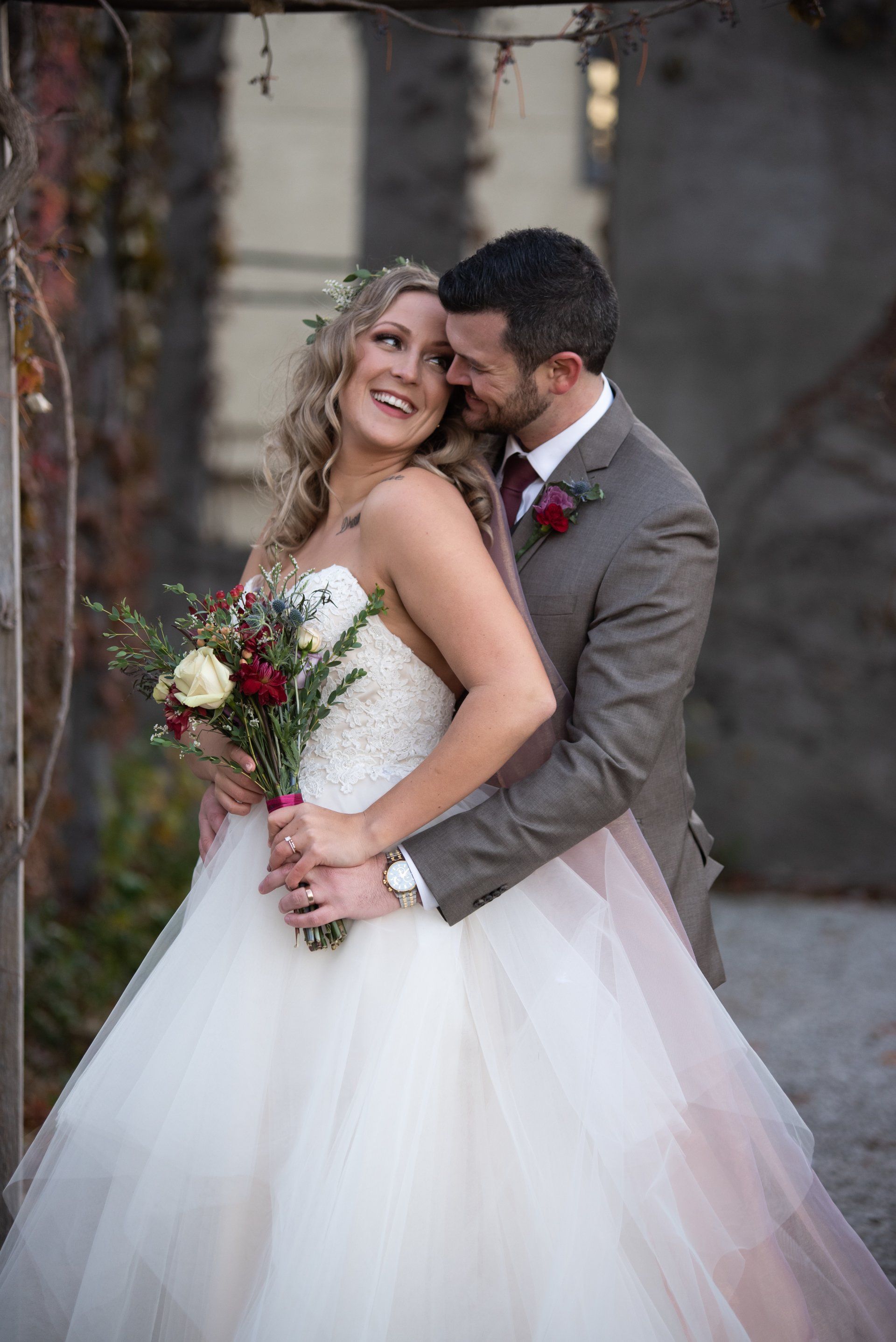 Bride and groom embracing, smiling. She wears a white wedding dress; he wears a gray suit. Outdoors.