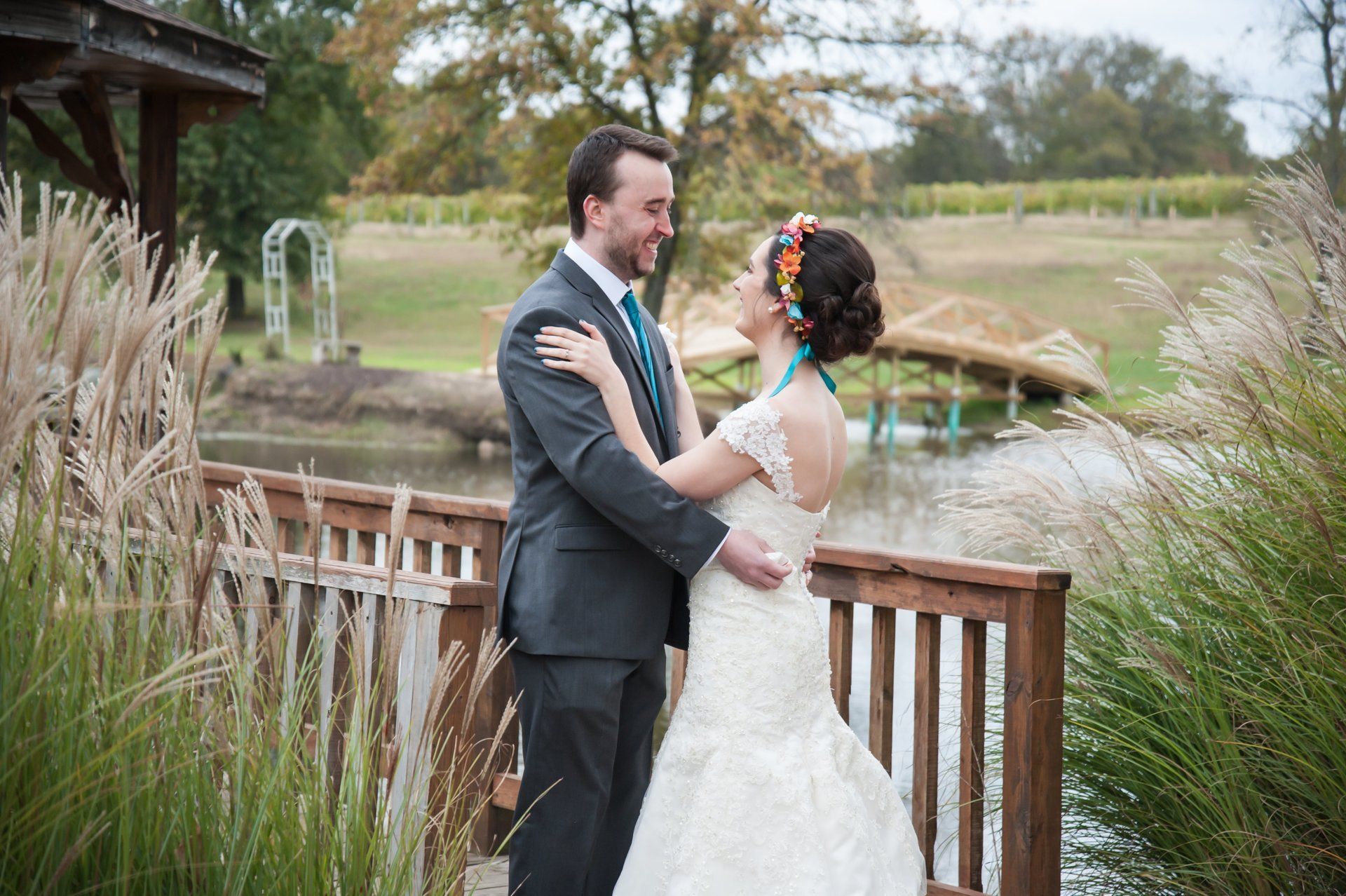 Bride and groom embrace on wooden deck, overlooking lake at outdoor wedding.