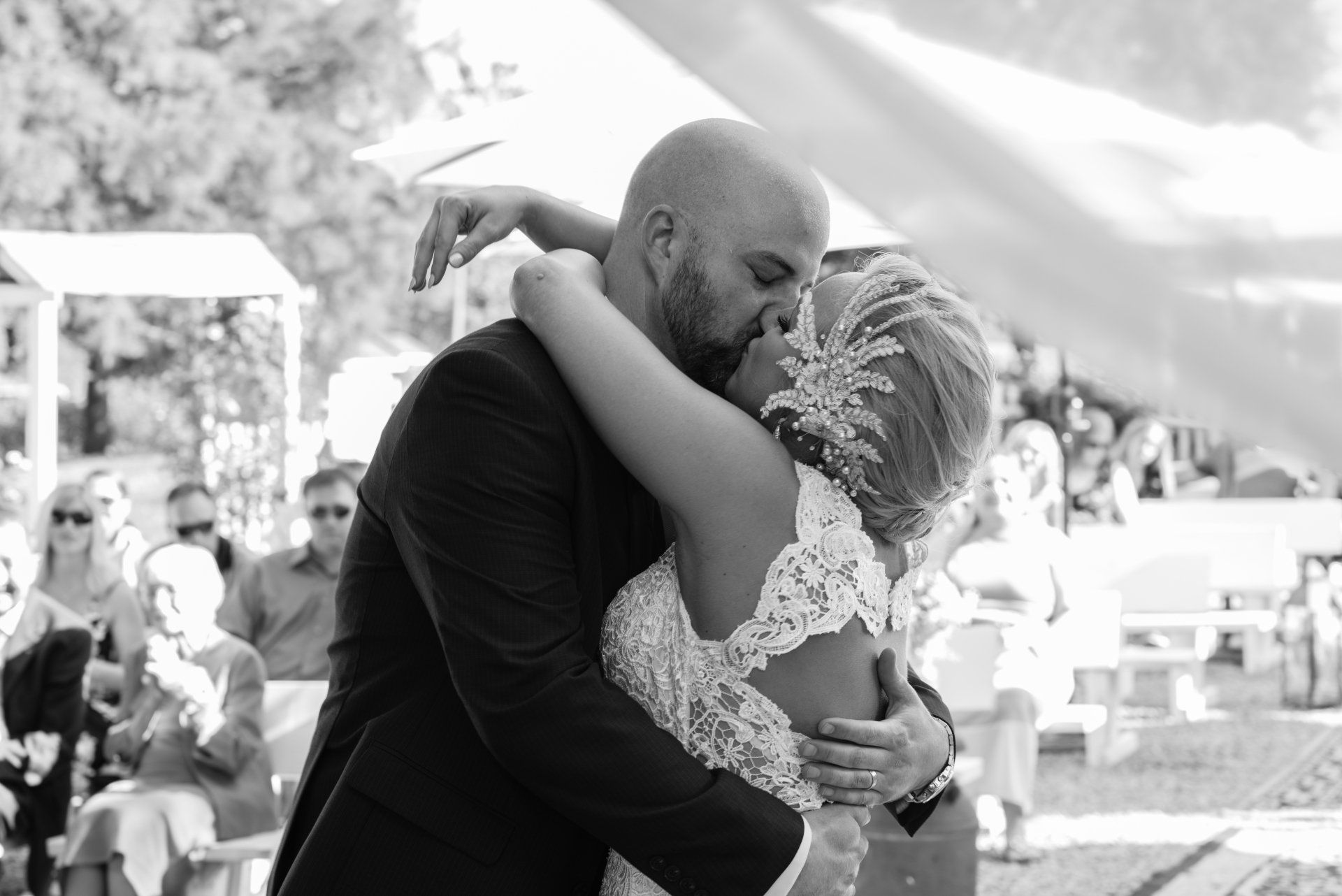 Newlyweds embrace and kiss passionately outdoors at wedding ceremony.