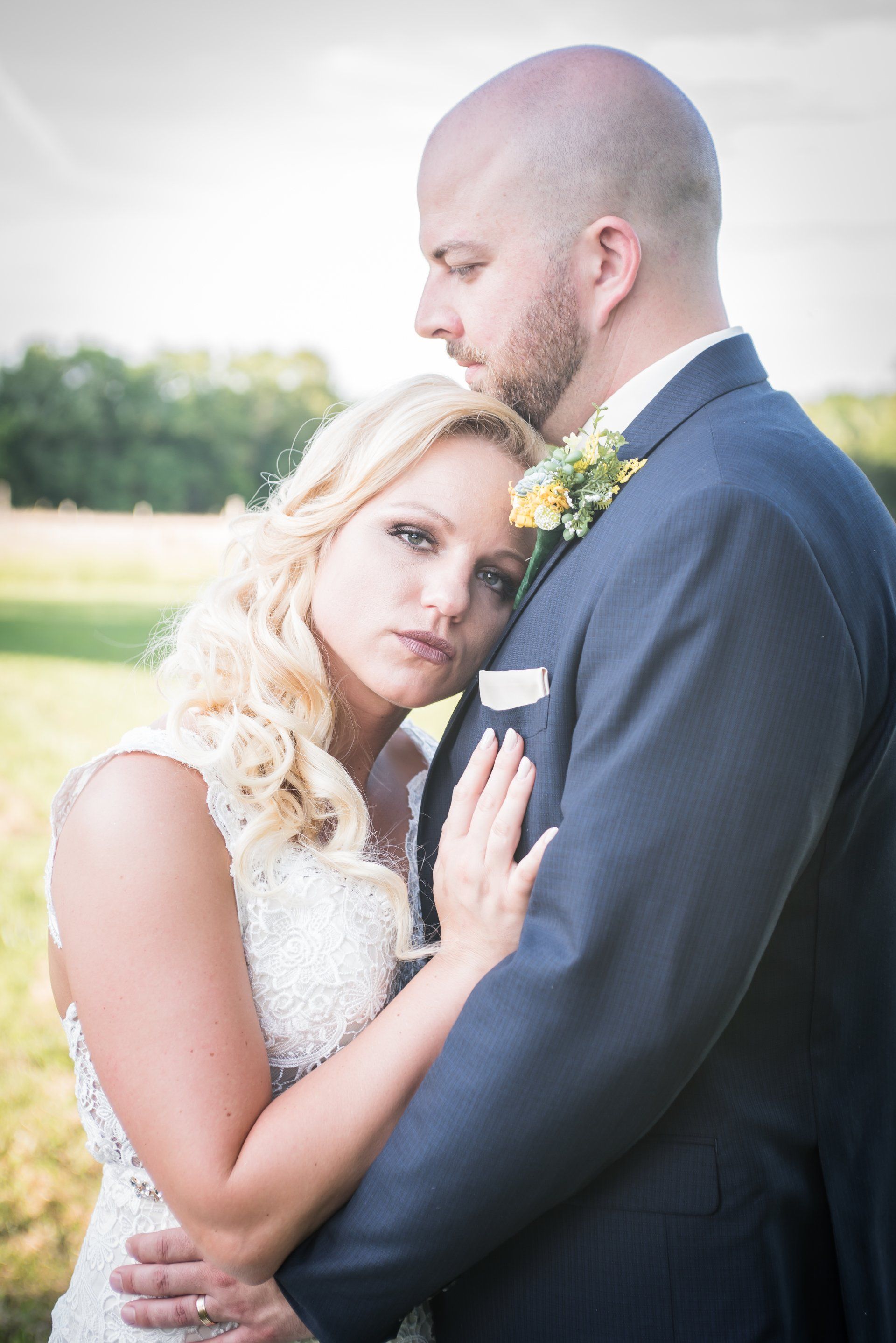 Bride and groom embrace outdoors; bride rests head on groom's chest. Groom wears blue suit; bride in white gown.