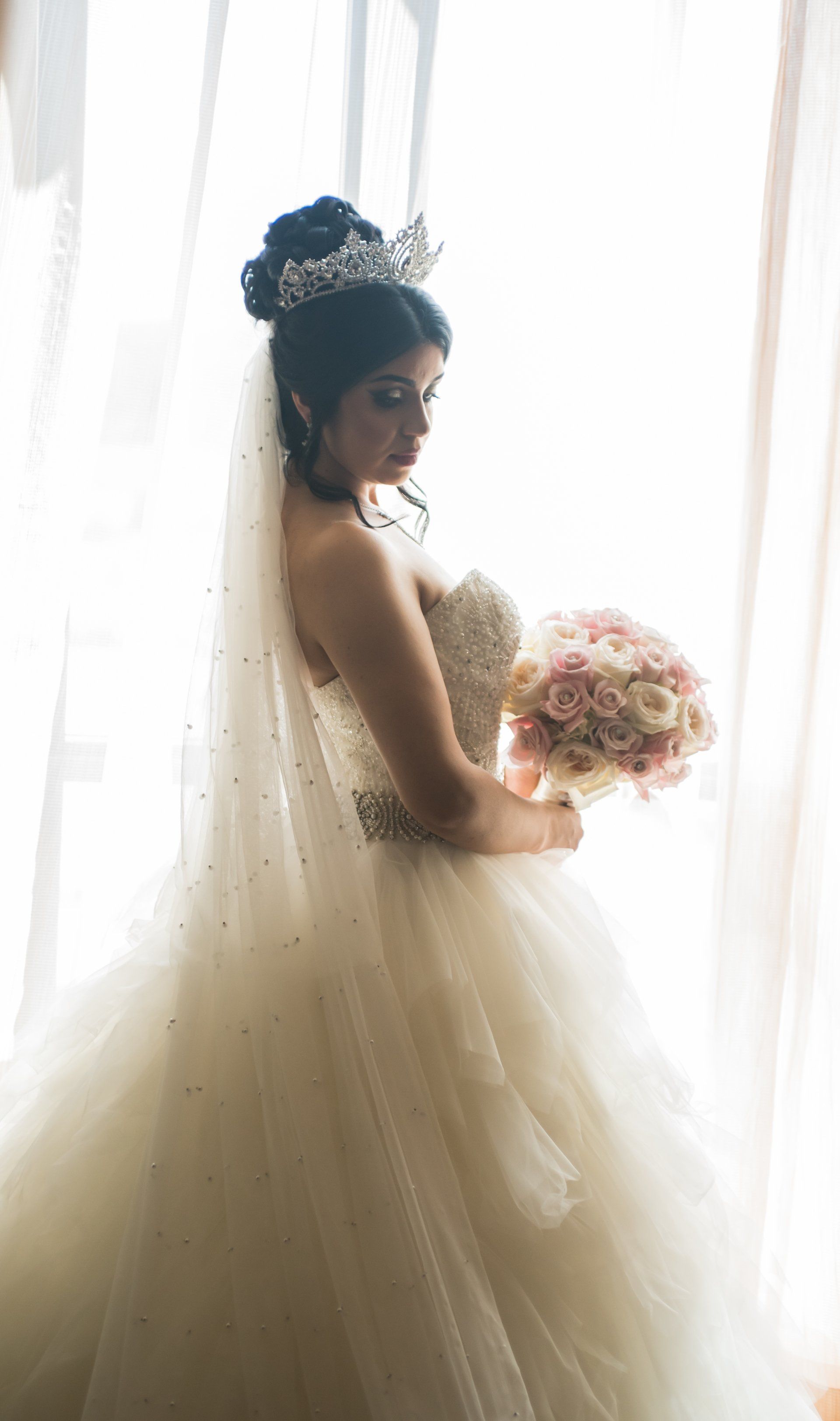 Bride in white gown and tiara holding pink bouquet, lit by a window.