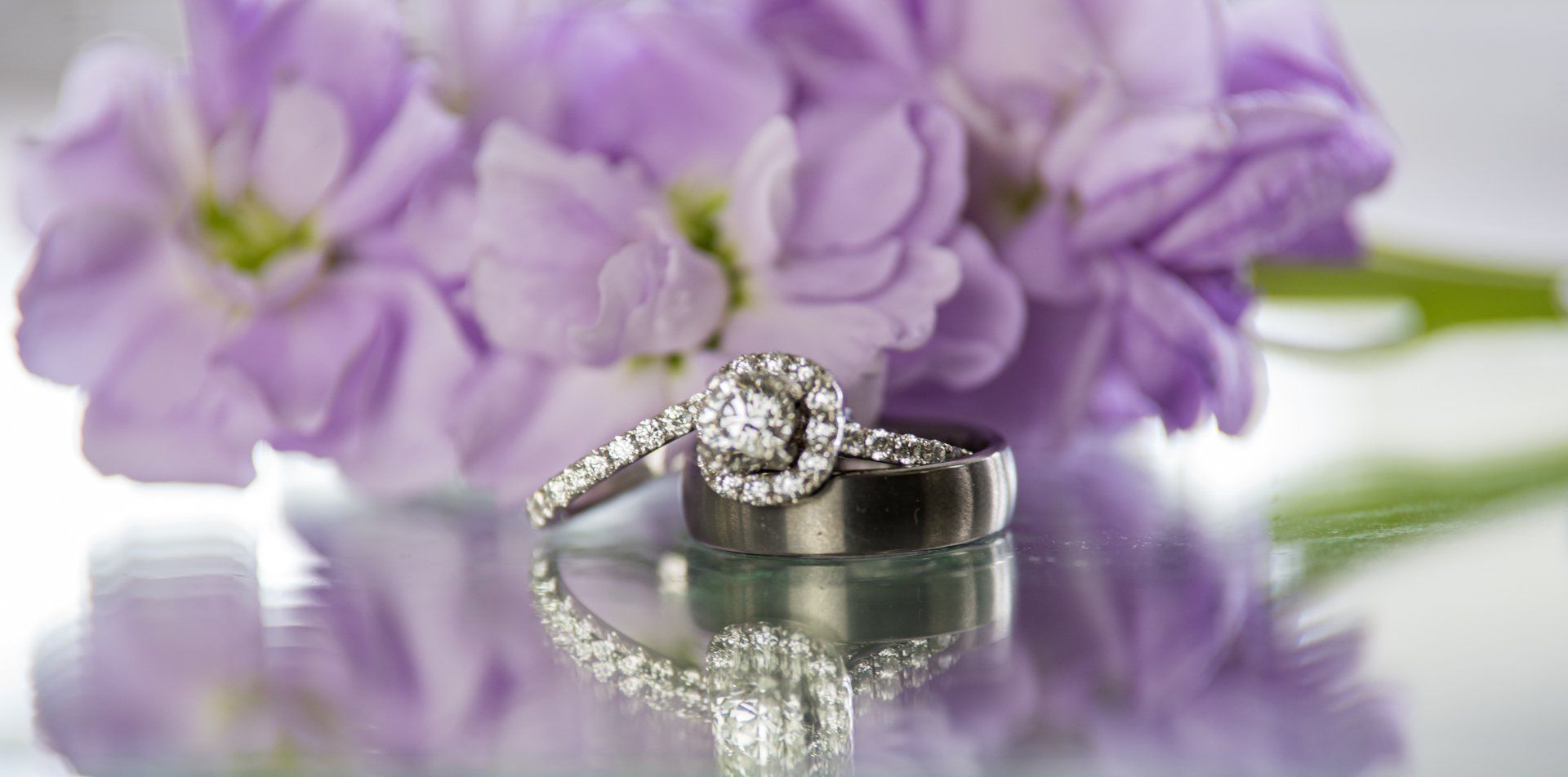 Two wedding rings with purple flowers in the background, reflected on a shiny surface.
