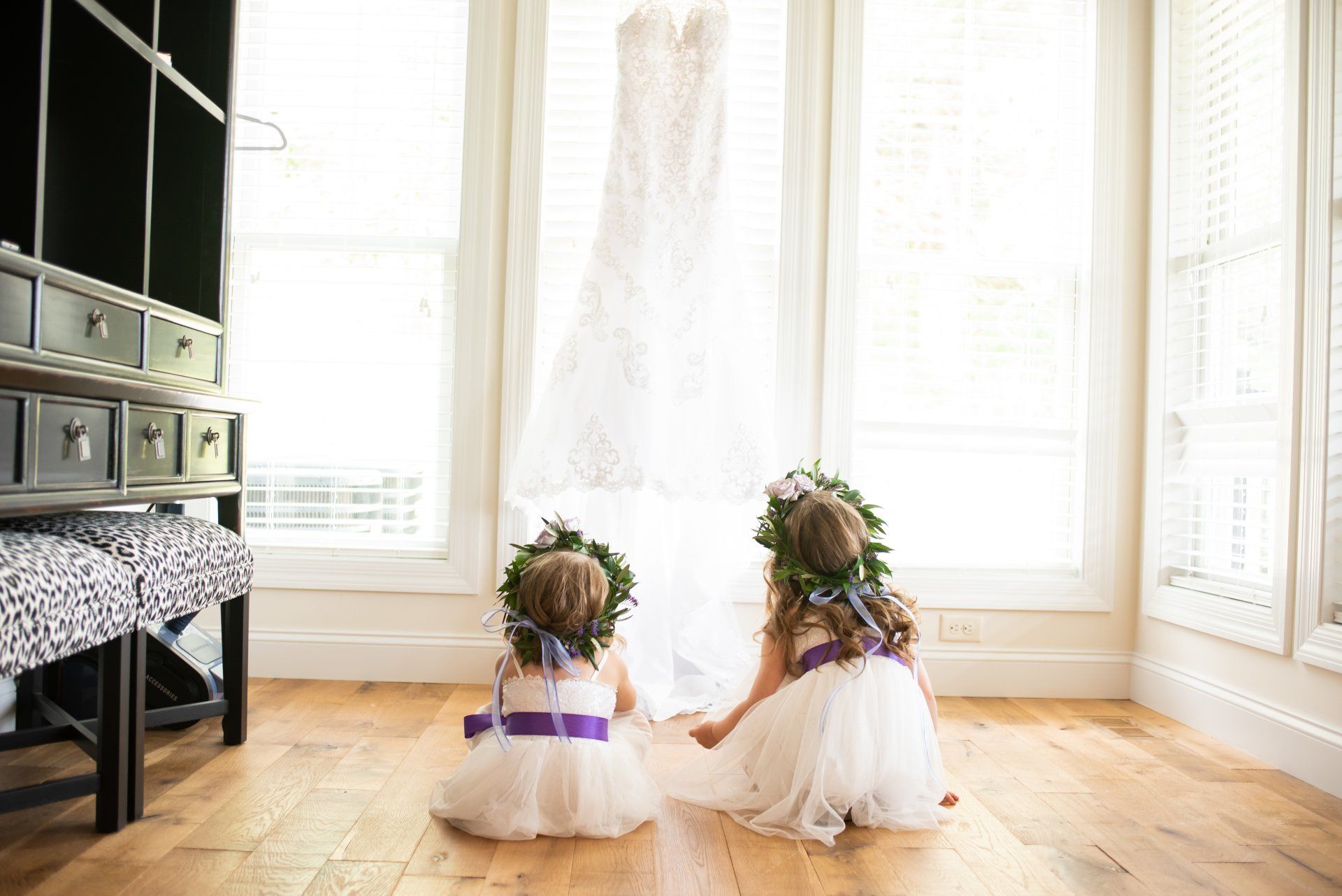 Two flower girls in white dresses with purple sashes and flower crowns, gazing at a wedding dress hanging in a window.