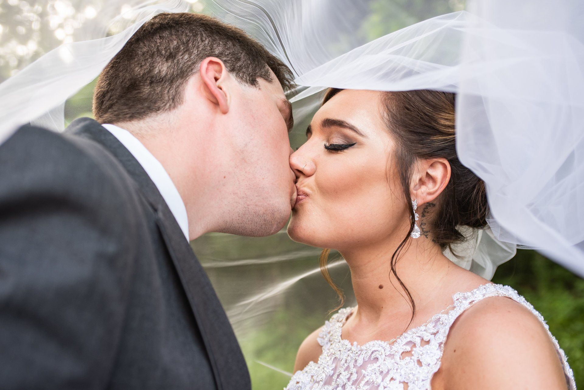 Couple kissing under a sheer white veil. The woman wears a white lace wedding dress and has her hair up.