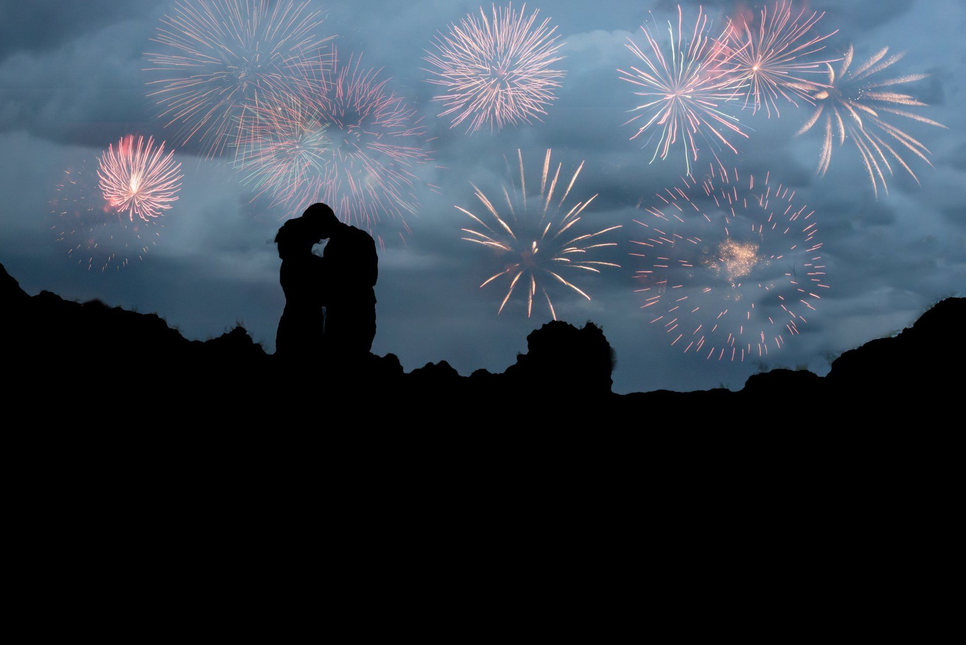 Silhouetted couple kissing with fireworks exploding overhead, against a dark sky.