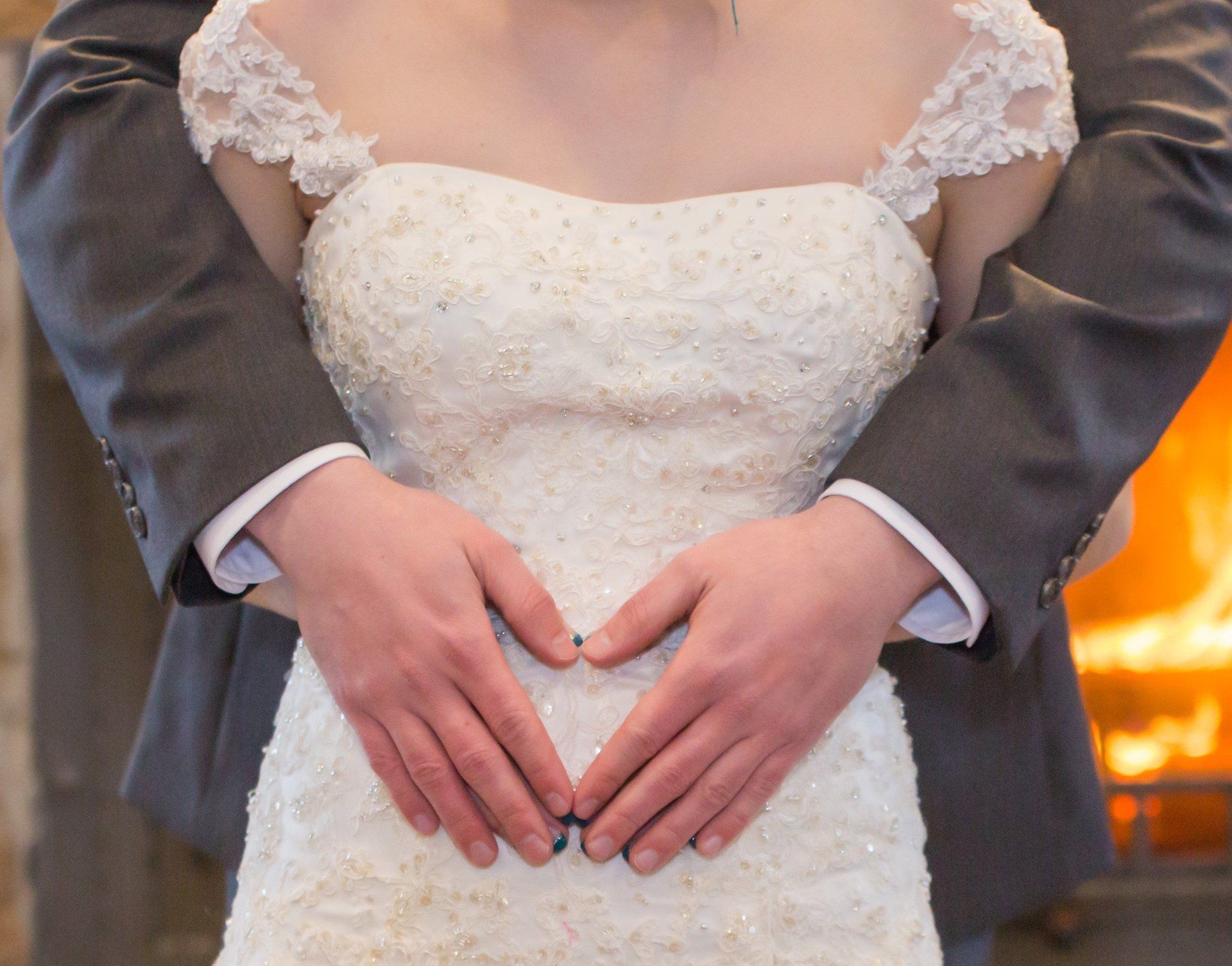 Groom's arms forming a heart around bride in white lace wedding dress, fireplace in background.