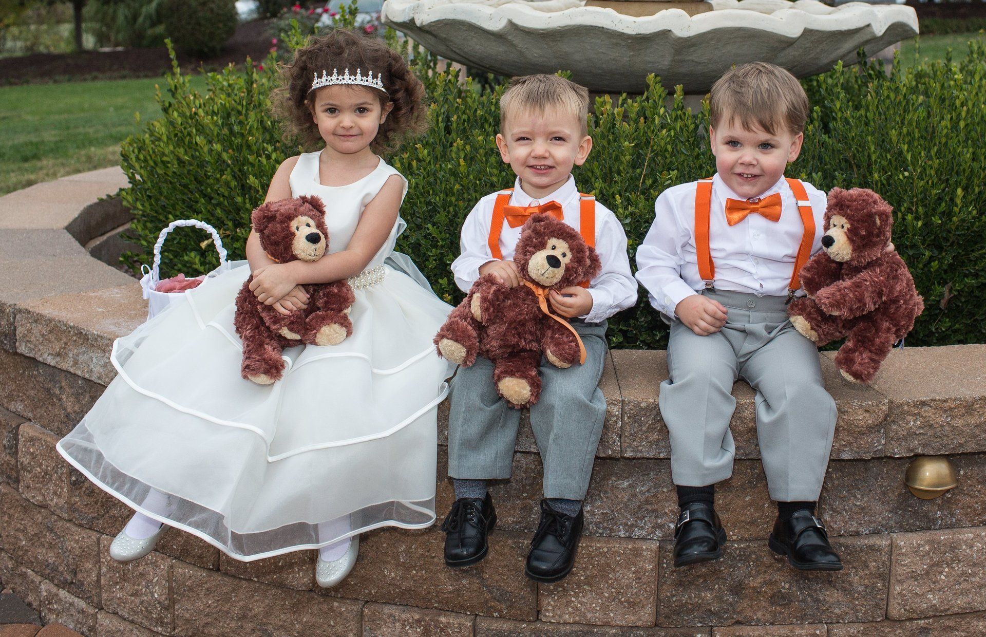 Three children in formal wear holding teddy bears sit on a stone wall.