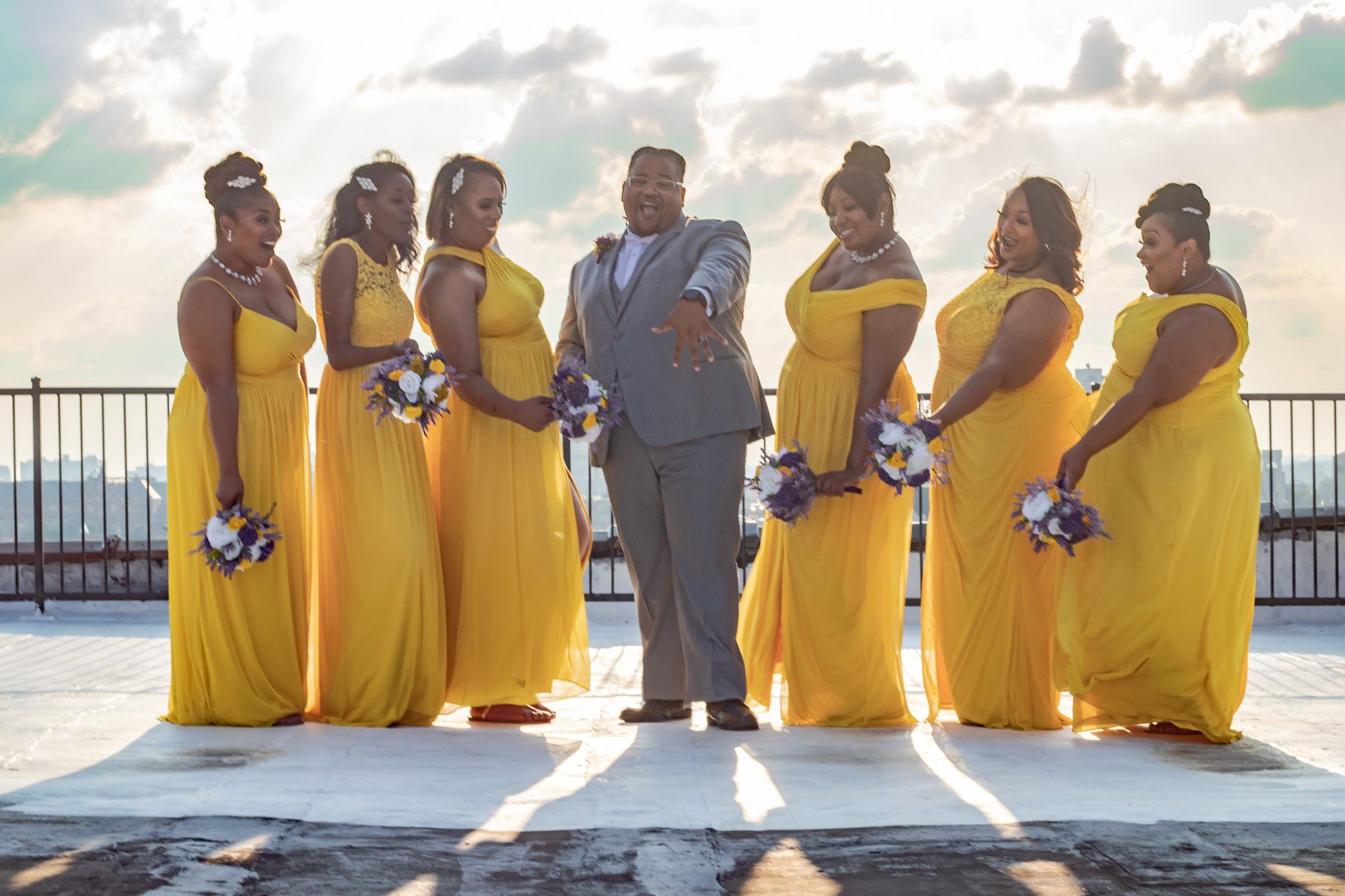 Groom with wedding party on a rooftop. Six bridesmaids in yellow dresses, groom in grey suit, sunlight.