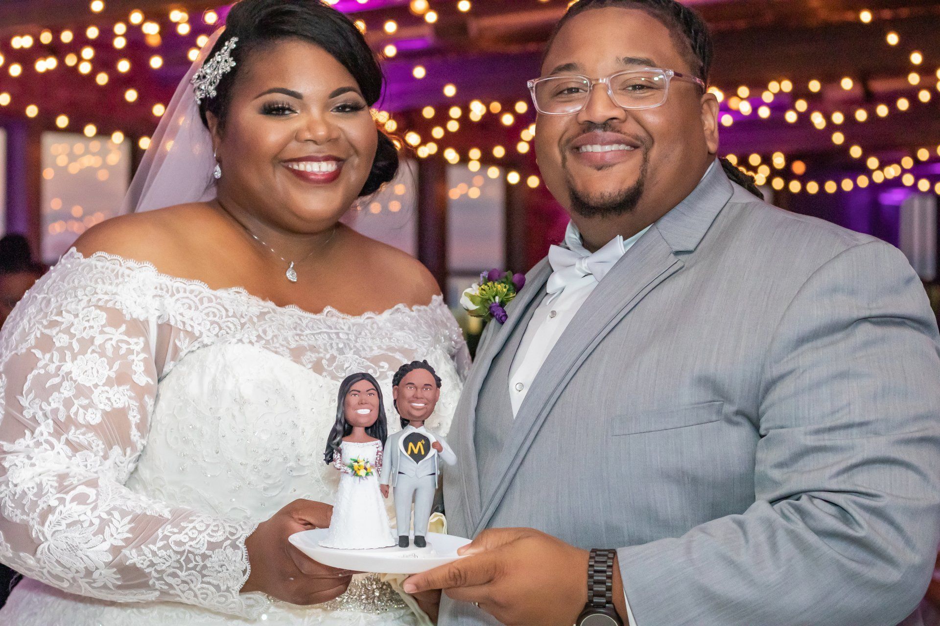 Newlyweds holding a wedding cake with a topper; the bride wears a lace gown, and the groom a suit, smiling.
