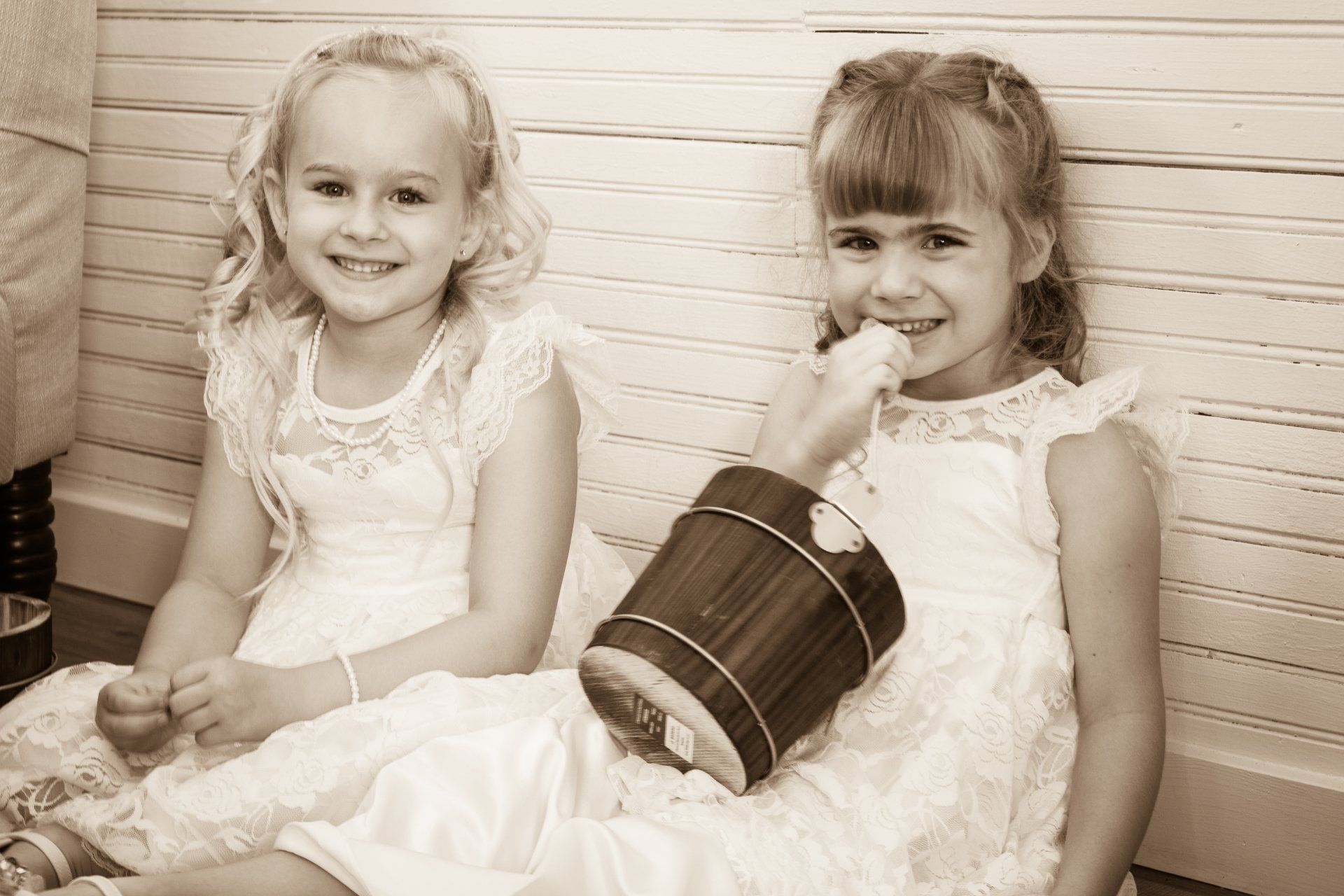 Two girls in white dresses smile, one holding a wooden bucket.