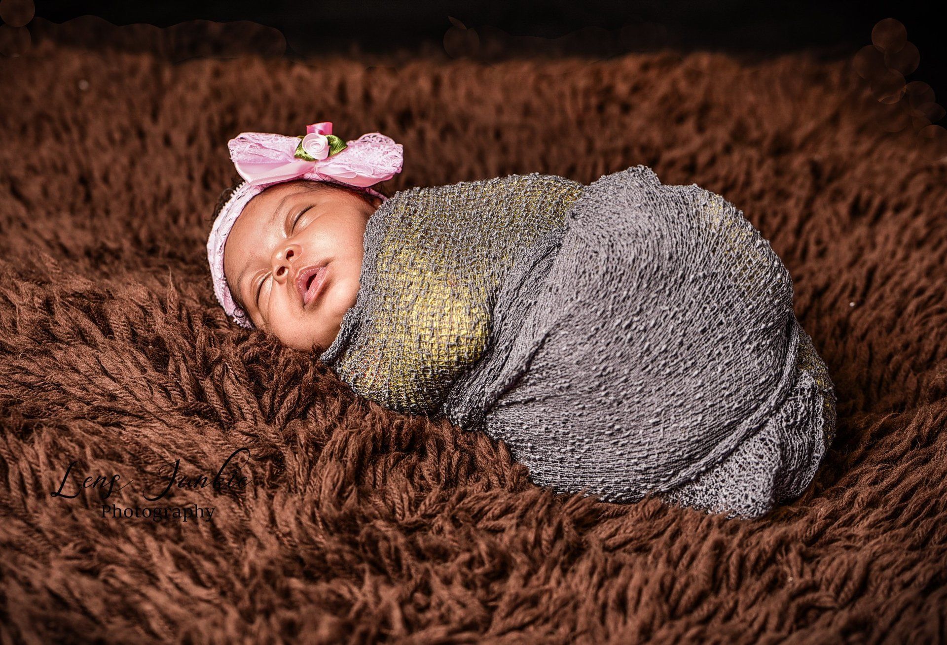 Newborn baby wrapped in shimmery fabric, wearing a pink headband with a flower, sleeping on a brown furry blanket.
