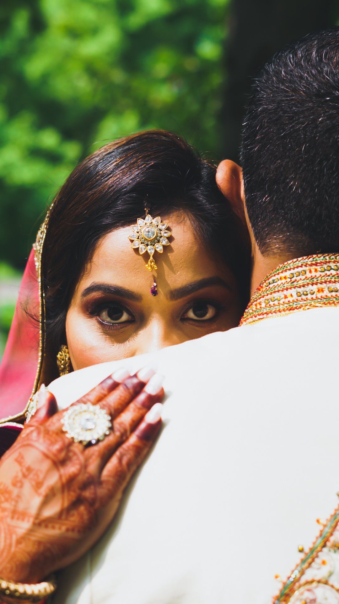 Woman in ornate headpiece looks over shoulder while embracing someone wearing a gold embroidered white garment.