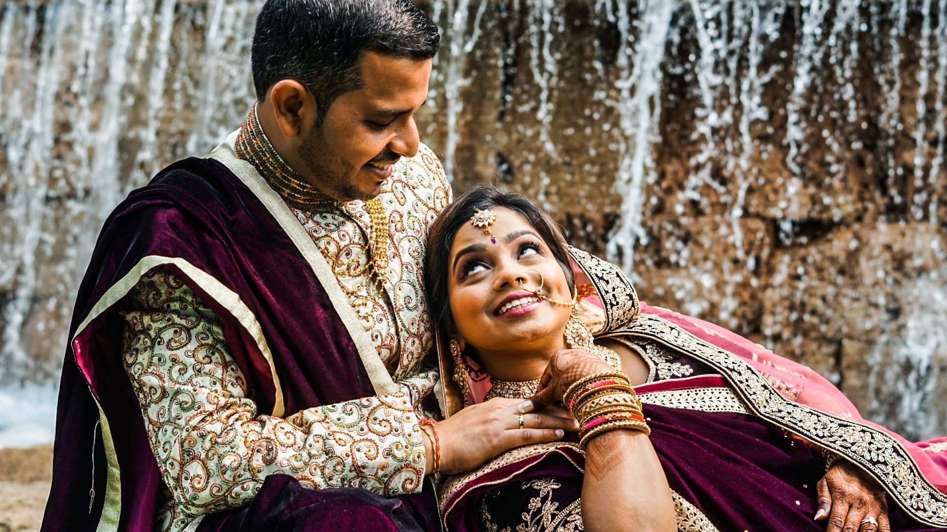Couple in formalwear by a waterfall. Man leans in, woman gazes up smiling.