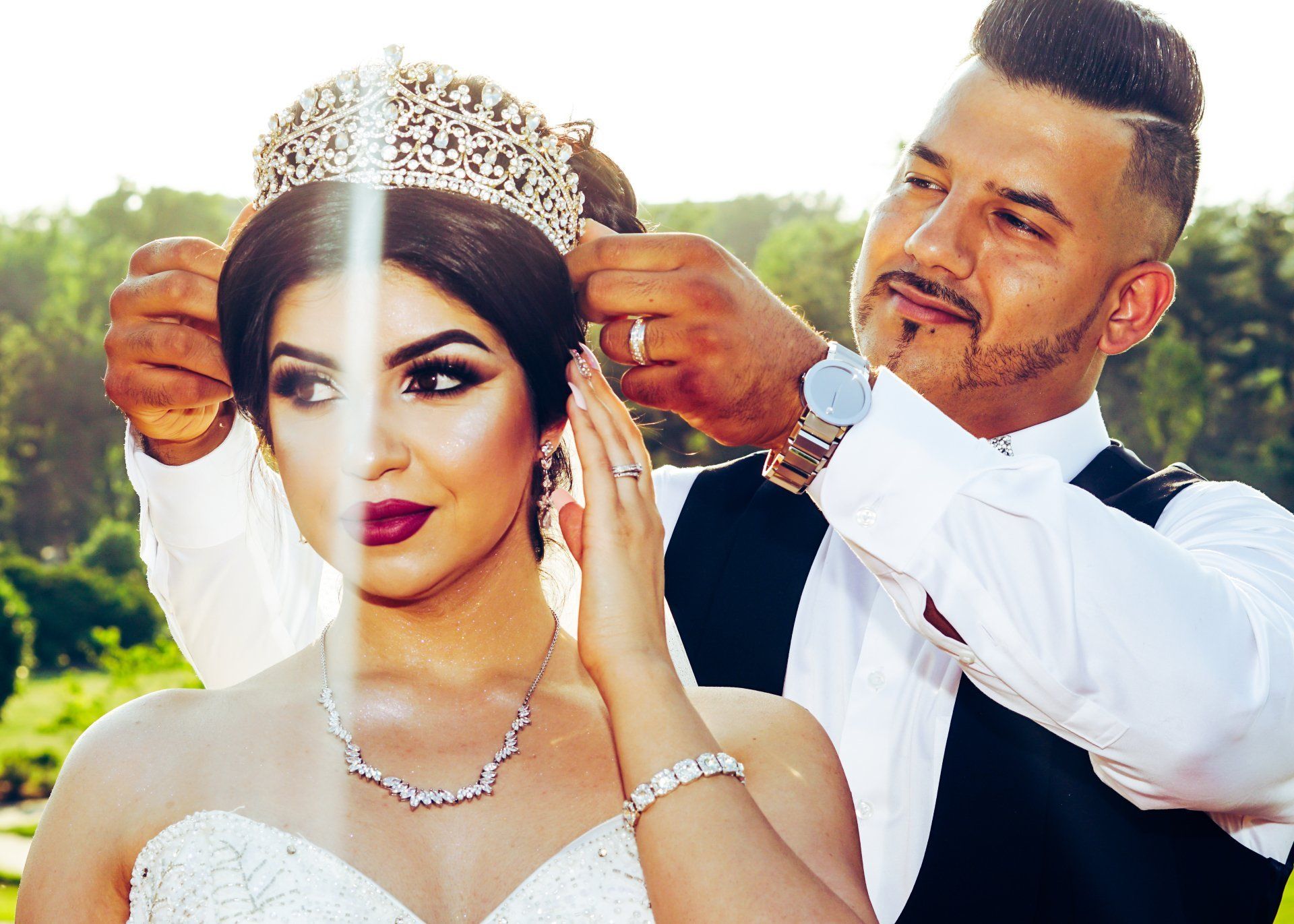 Groom places a jeweled crown on bride's head, outdoors. Both wear formal attire; bride has elegant makeup.