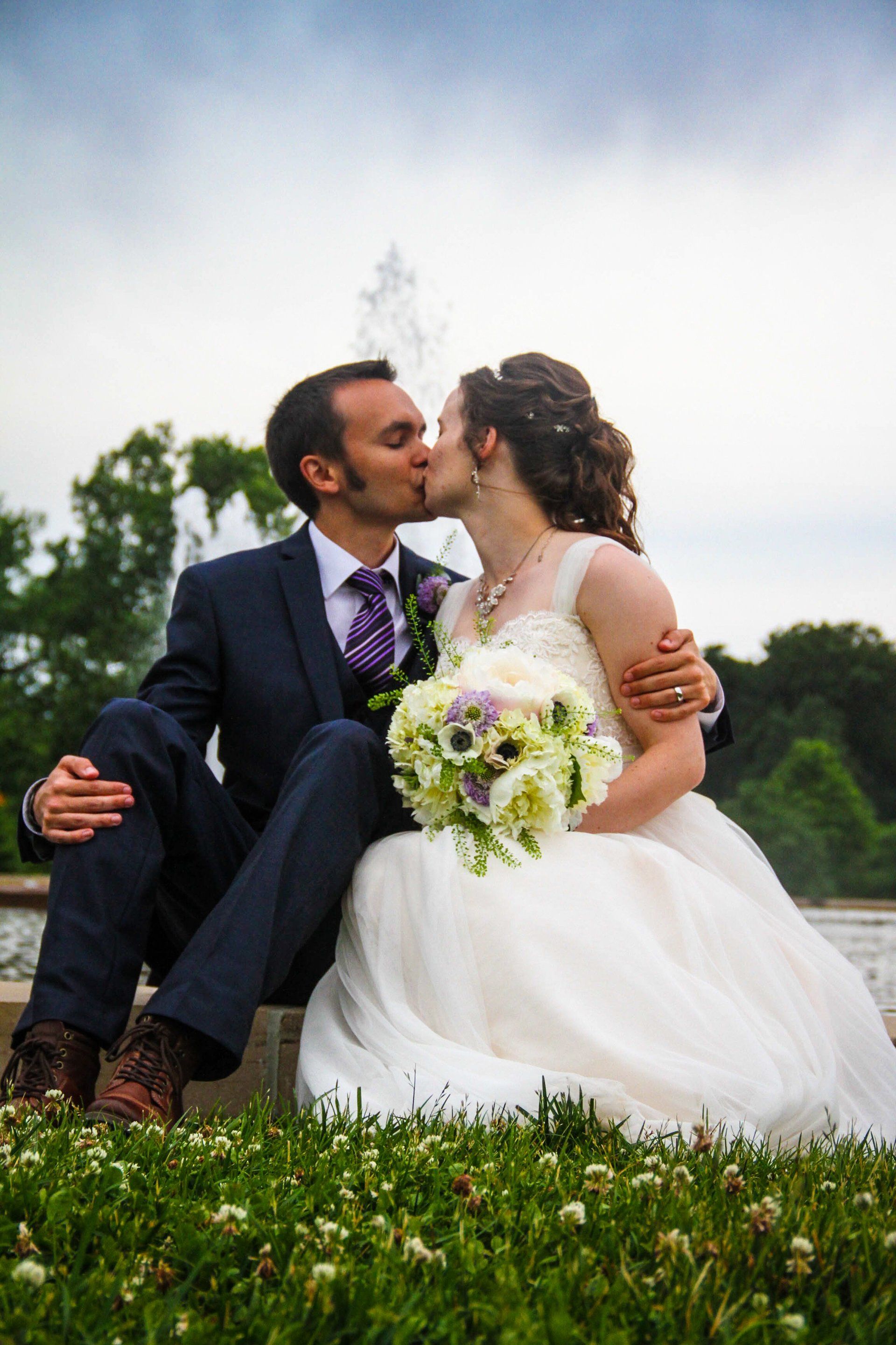 Couple kissing; man in blue suit, woman in white dress, seated near fountain.