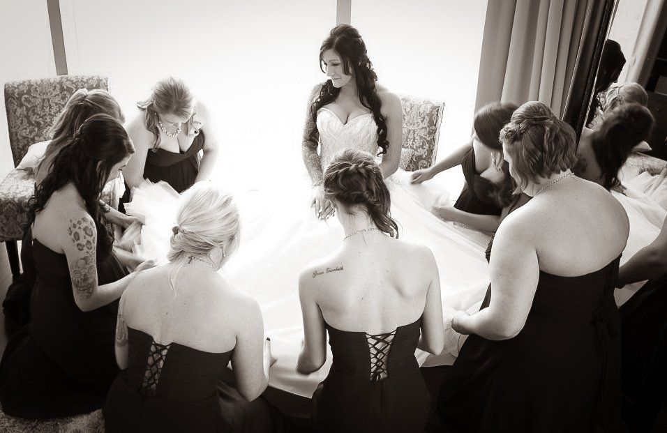 Bride in white dress surrounded by bridesmaids in black dresses, indoors, preparing for wedding.