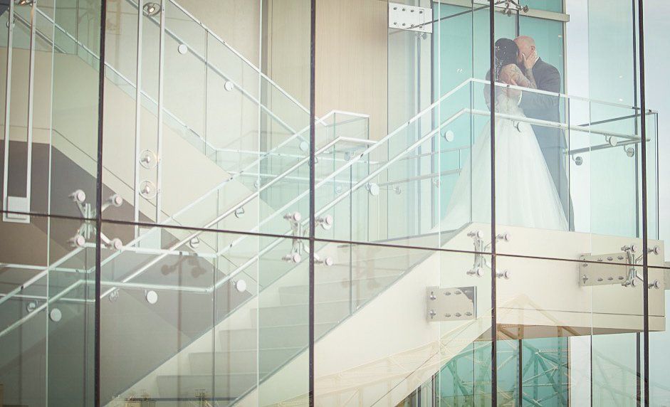 Couple kissing on a modern glass staircase, wedding attire, natural light.