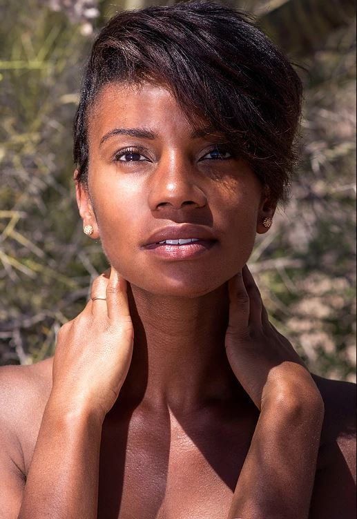 Woman with short dark hair holds her neck outdoors, looking at the camera with a calm expression.