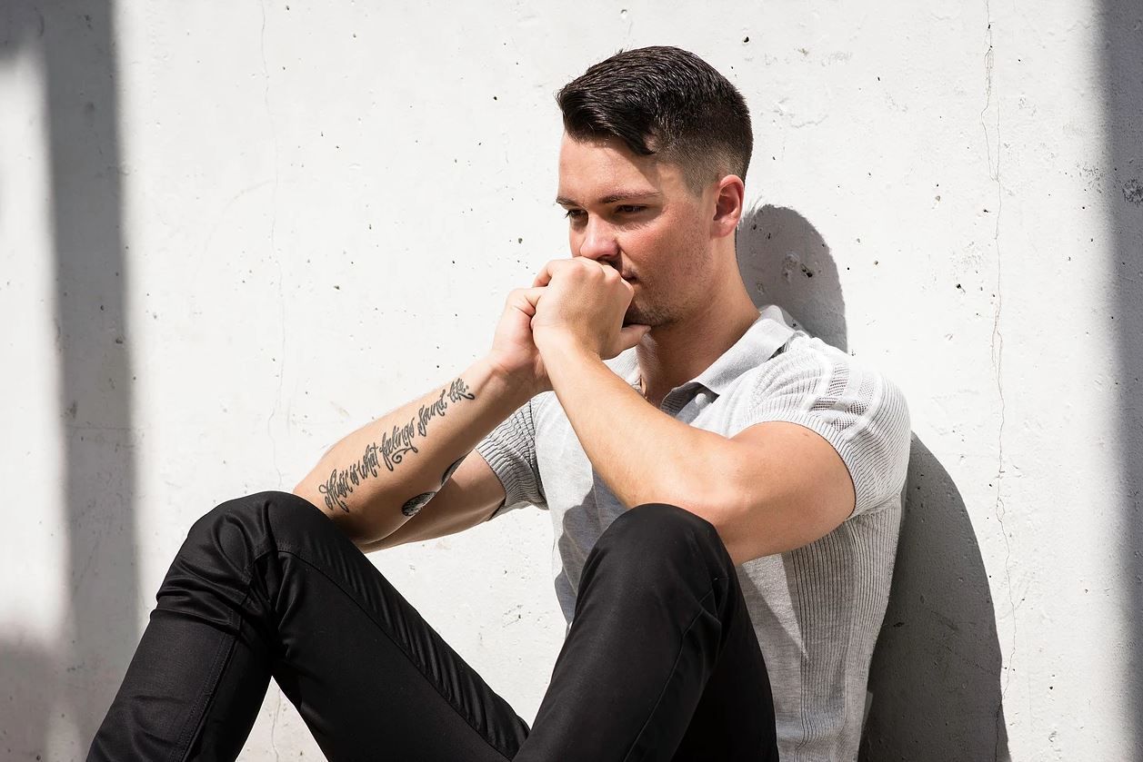 Man sitting with hands near his face, leaning against a white wall; possibly deep in thought.