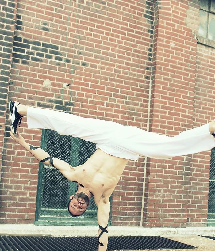 Man balancing in a handstand split, shirtless, against a brick wall.